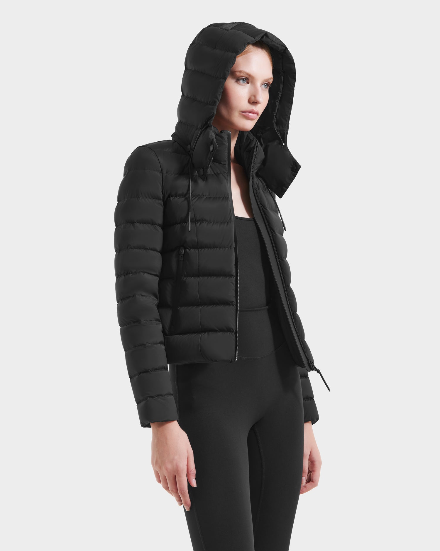 A woman wearing the Rudsak MELISSA - 8125054 Black fitted puffer jacket with a hood, paired with black leggings and a top, stands against a plain white background.