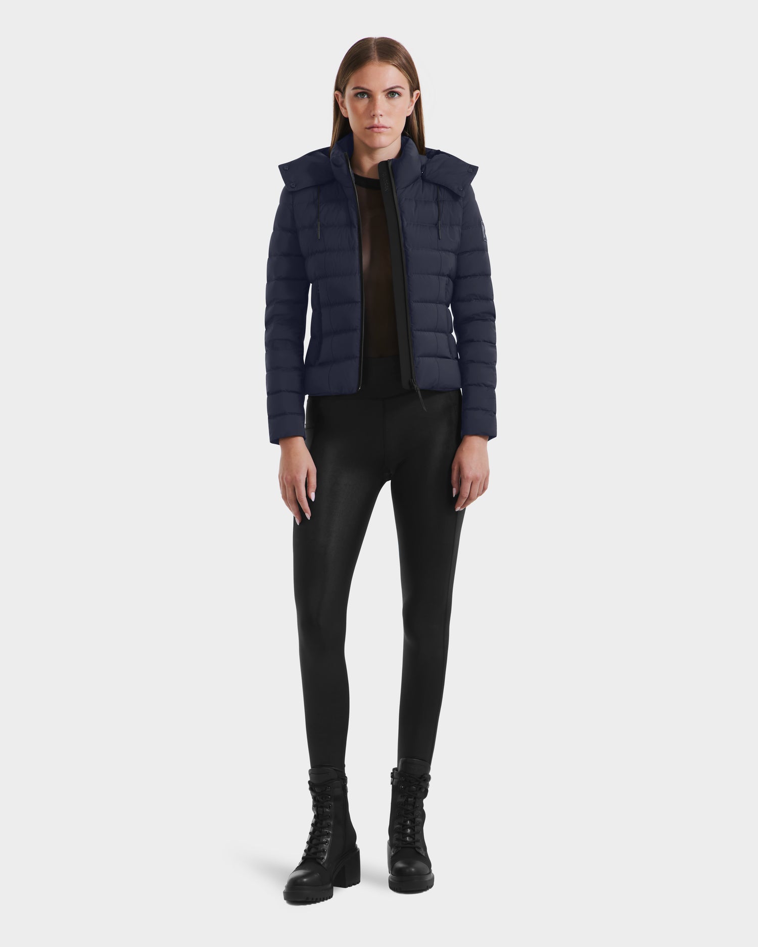 A woman faces forward in the Rudsak MELISSA - 8125054 Navy womens puffer jacket, styled over a black sheer top, black leggings, and black lace-up boots against a plain white background.
