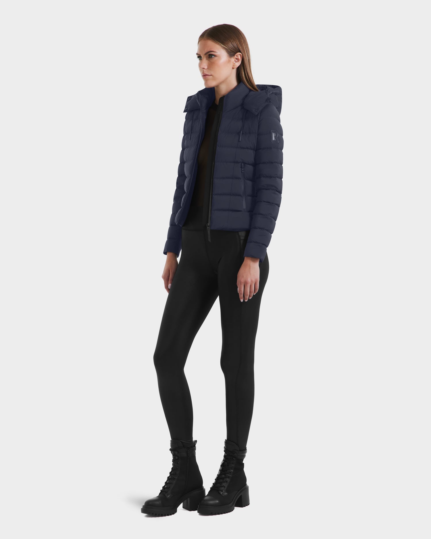 A woman stands slightly turned to the side, wearing the Rudsak MELISSA - 8125054 Navy womens puffer jacket with RDS certified down, paired with black leggings and lace-up boots, set against a white background.