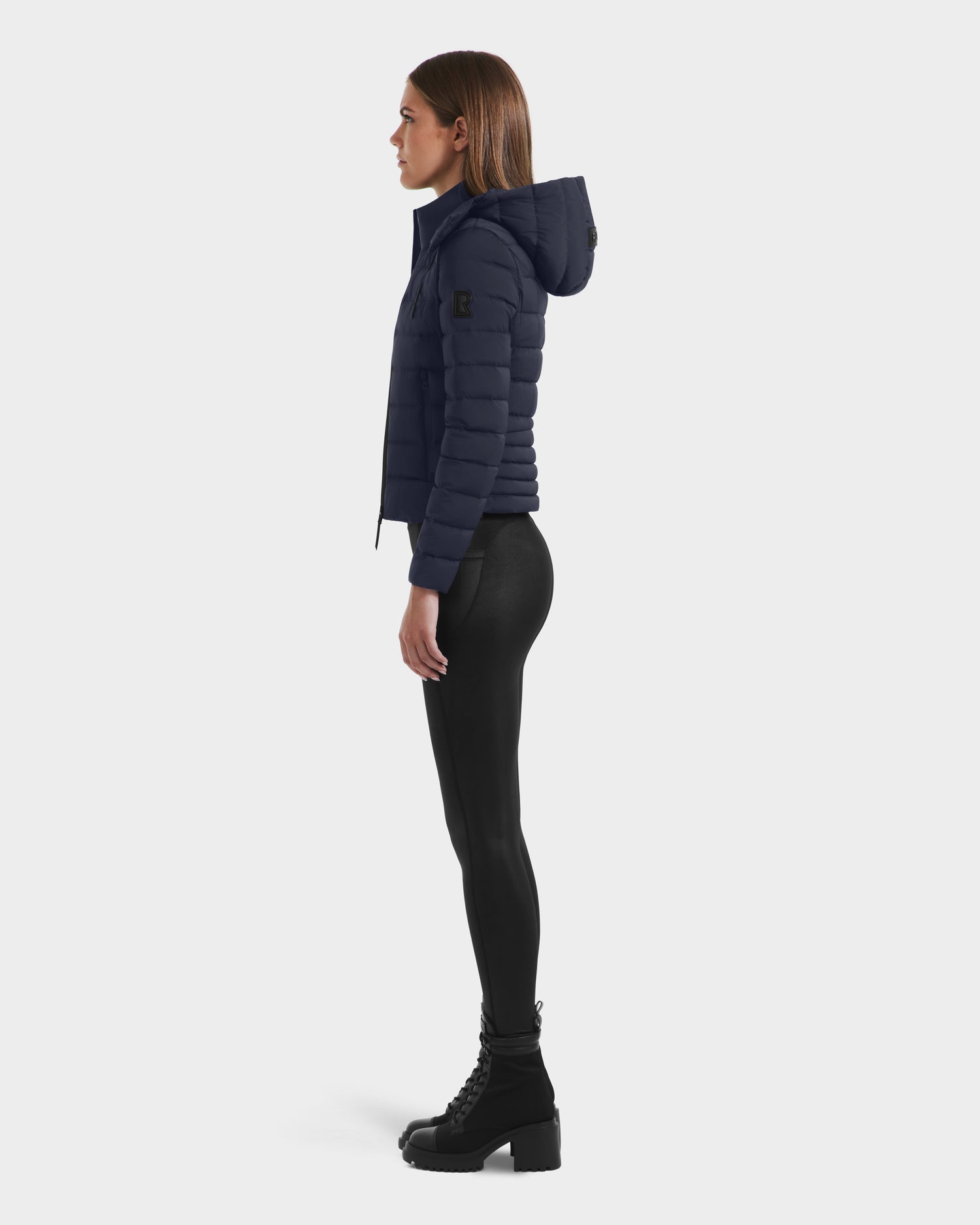 A woman stands in profile wearing the Rudsak MELISSA - 8125054 Navy RDS certified down puffer jacket with a hood, paired with black leggings and lace-up boots against a plain white background.