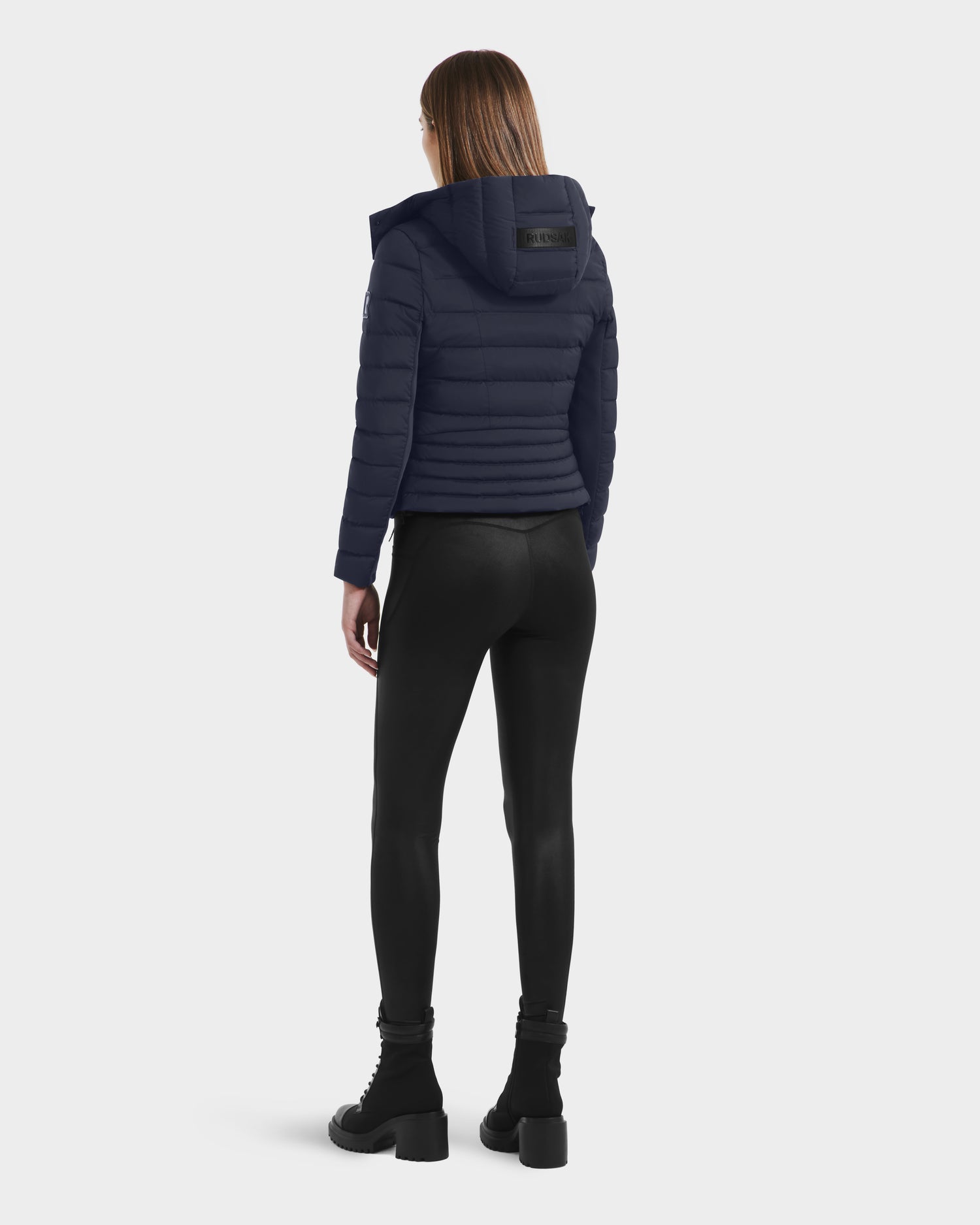A person stands facing away, wearing the Rudsak MELISSA - 8125054 Navy womens puffer jacket with a hood, black leggings, and black heeled boots against a plain light background.
