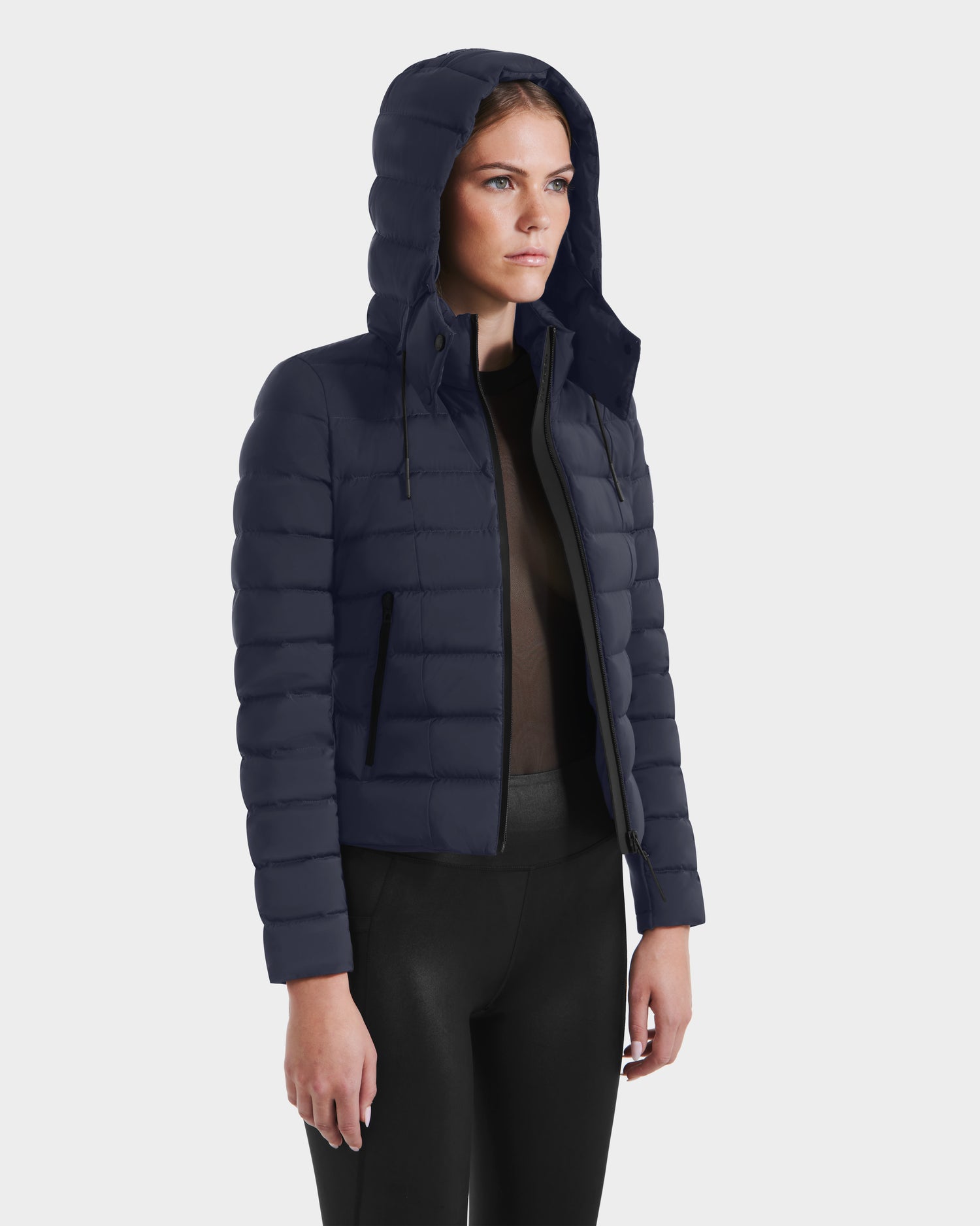 A person models the Rudsak MELISSA - 8125054 Navy womens puffer jacket with a hood, black pants, and a dark top, standing at an angle against a plain light background. This lightweight coat uses RDS certified down.