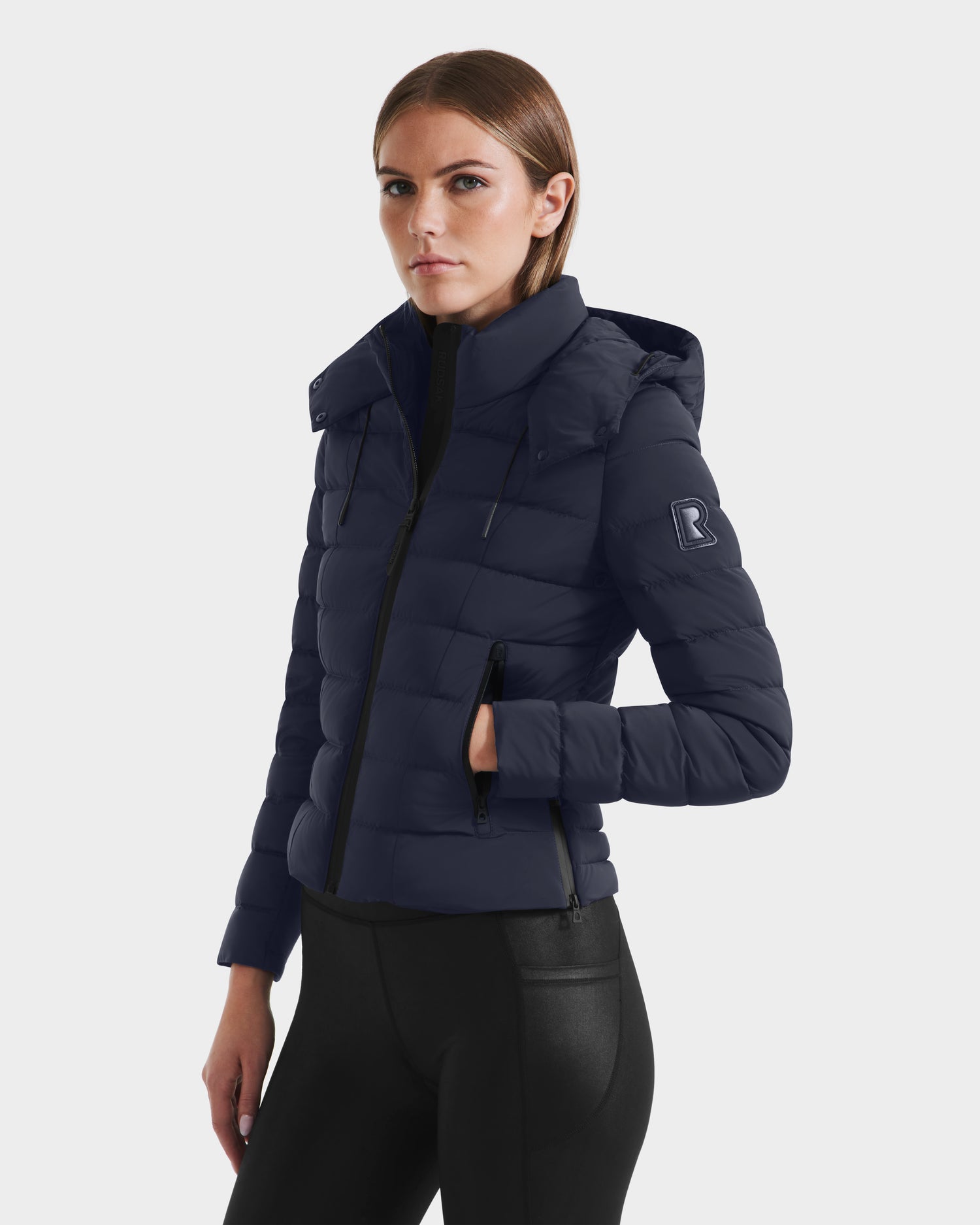 A woman wearing the MELISSA - 8125054 Navy puffer jacket by Rudsak, made with RDS certified down and a B logo on the sleeve, stands against a light background, looking at the camera with a neutral expression.