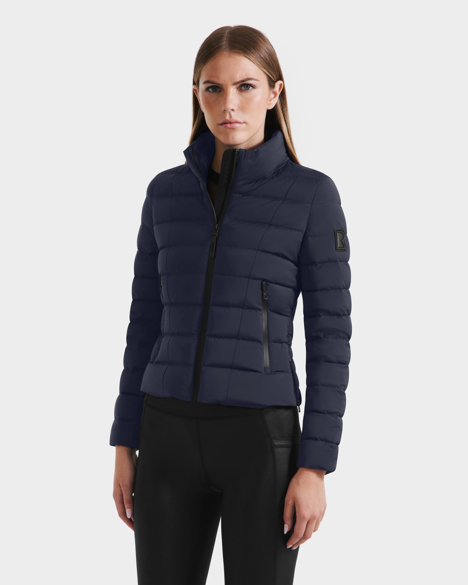 A woman with straight brown hair wears the Rudsak MELISSA - 8125054 Navy puffer jacket, featuring a fitted shape, high collar, and black zippered pockets, paired with black pants against a light background.