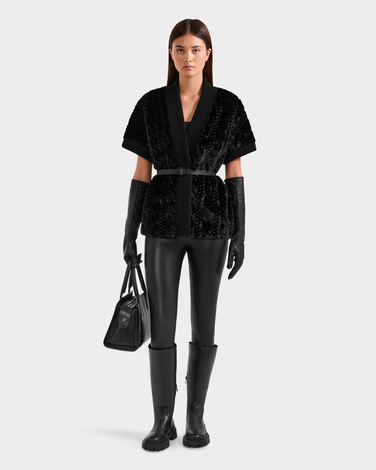 A woman faces forward in a Rudsak MENDOTA F - 8525561 black textured short-sleeve belted jacket, paired with black leather gloves, leather pants, knee-high boots, and holding a black handbag against a plain white background.