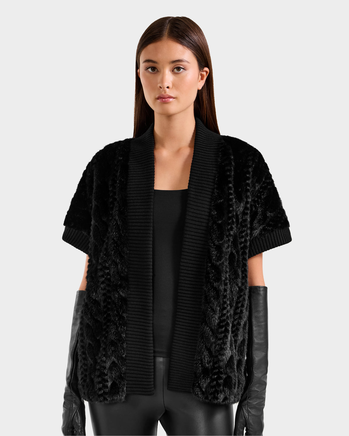 A woman with straight brown hair wears the Rudsak MENDOTA F - 8525561 Black textured short-sleeve cardigan over a black top, styled with black leather gloves and pants, posing against a plain light gray background.