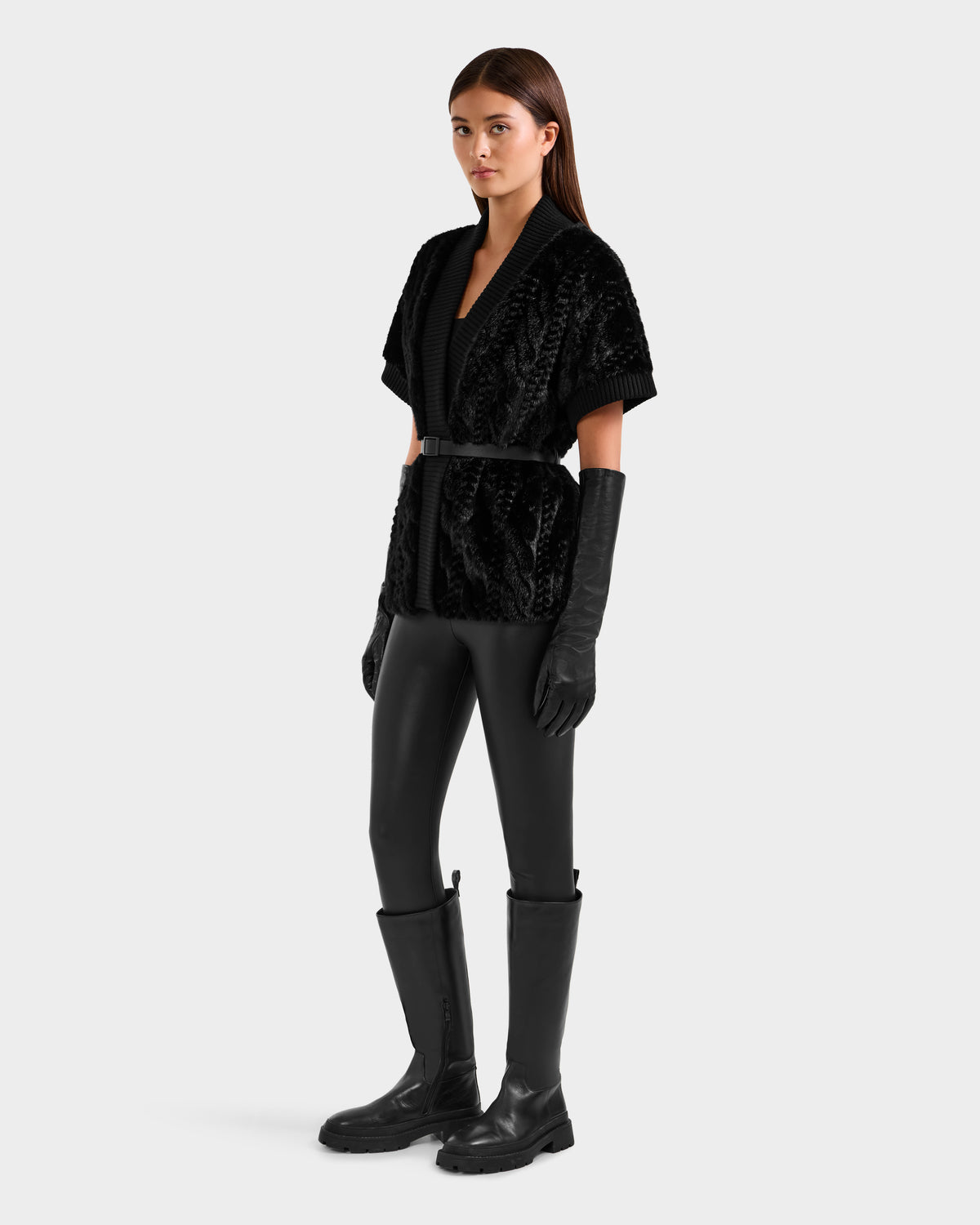 A woman wears the Rudsak MENDOTA F - 8525561 Black textured short-sleeve jacket with a belt, paired with black gloves, leather pants, and knee-high boots against a plain light backdrop.