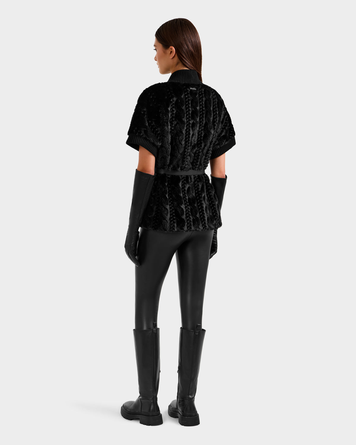A woman with straight brown hair stands facing away against a light gray background, wearing the Rudsak MENDOTA F - 8525561 Black textured short-sleeve jacket, black gloves, fitted pants, and knee-high boots.