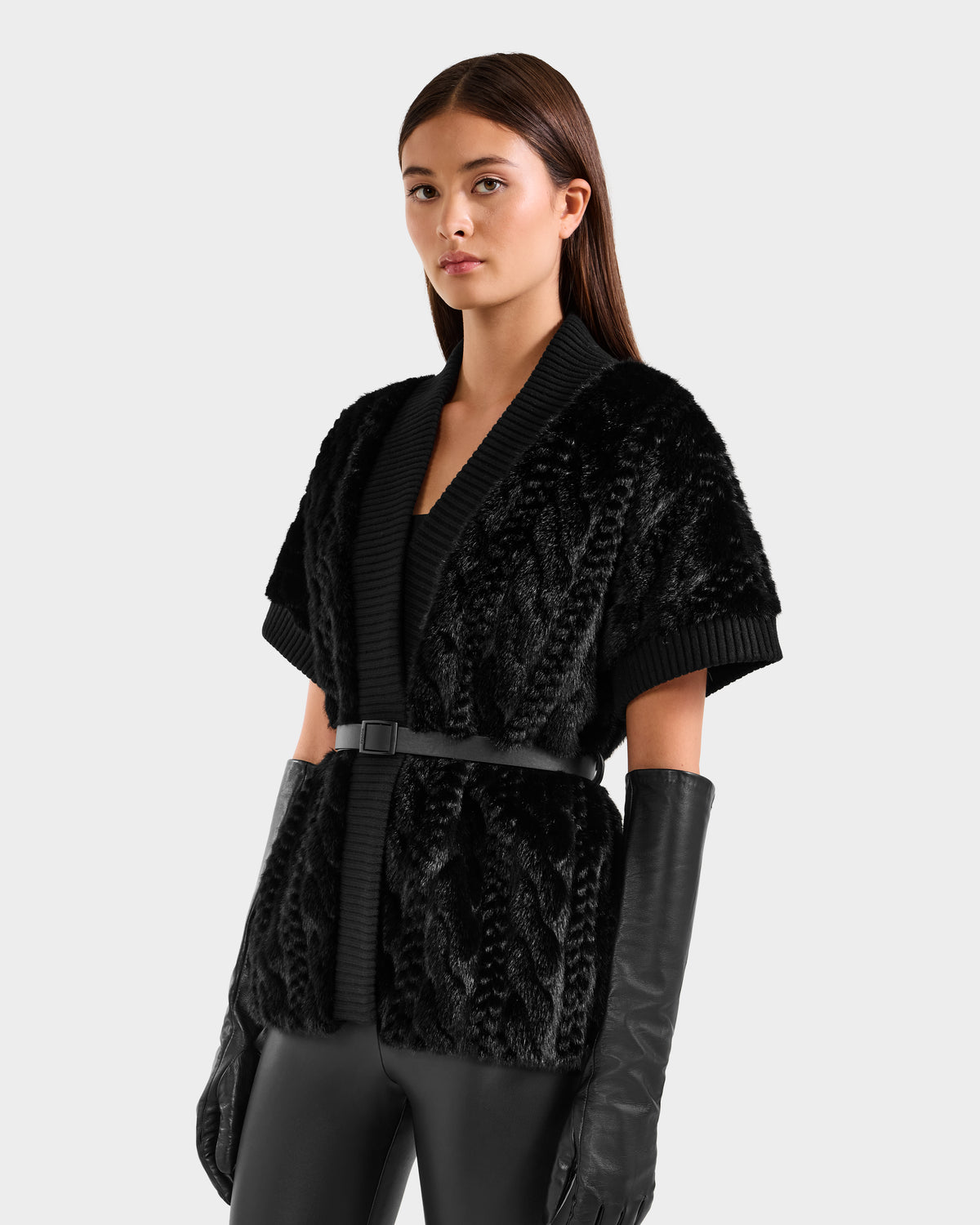 A woman with straight brown hair wears the Rudsak MENDOTA F - 8525561 Black short-sleeve cardigan, styled with a vegan leather belt, black long gloves, and pants, standing against a plain light background.