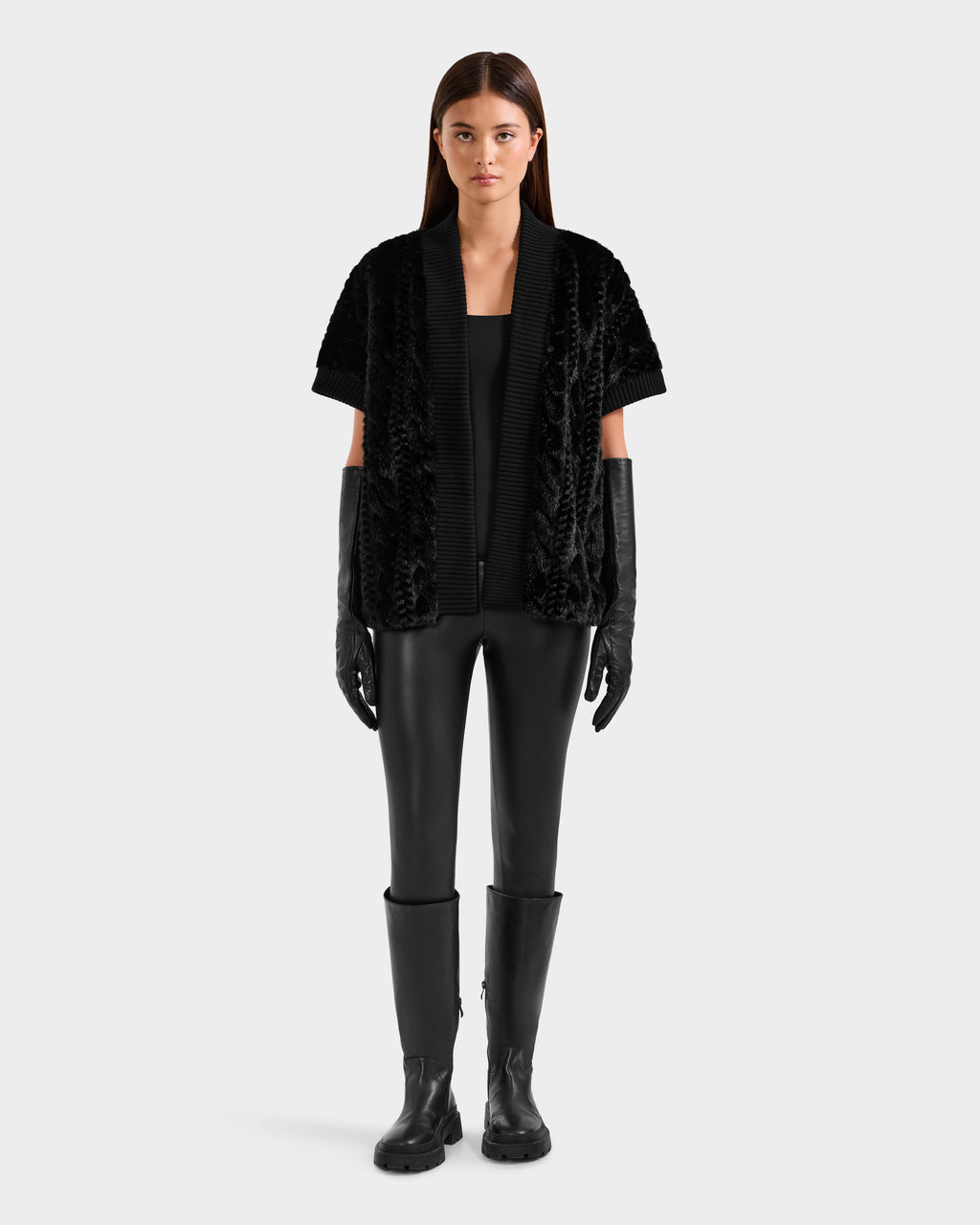 A woman faces forward in the Rudsak MENDOTA F - 8525561 Black jacket, styled with black faux fur top, leather pants, long black gloves, and knee-high boots against a plain light gray background.
