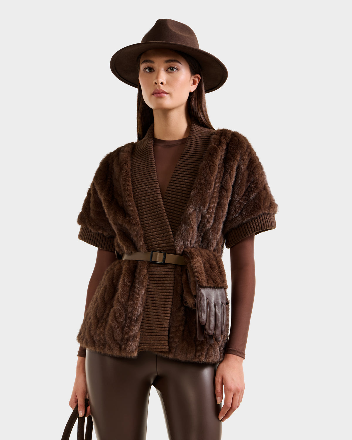 A woman wears the Rudsak MENDOTA F - 8525561 Mocha textured faux fur cardigan with vegan leather belt, brown gloves at her waist, a long-sleeve wrap top, and brown leather pants, standing against a light gray background.