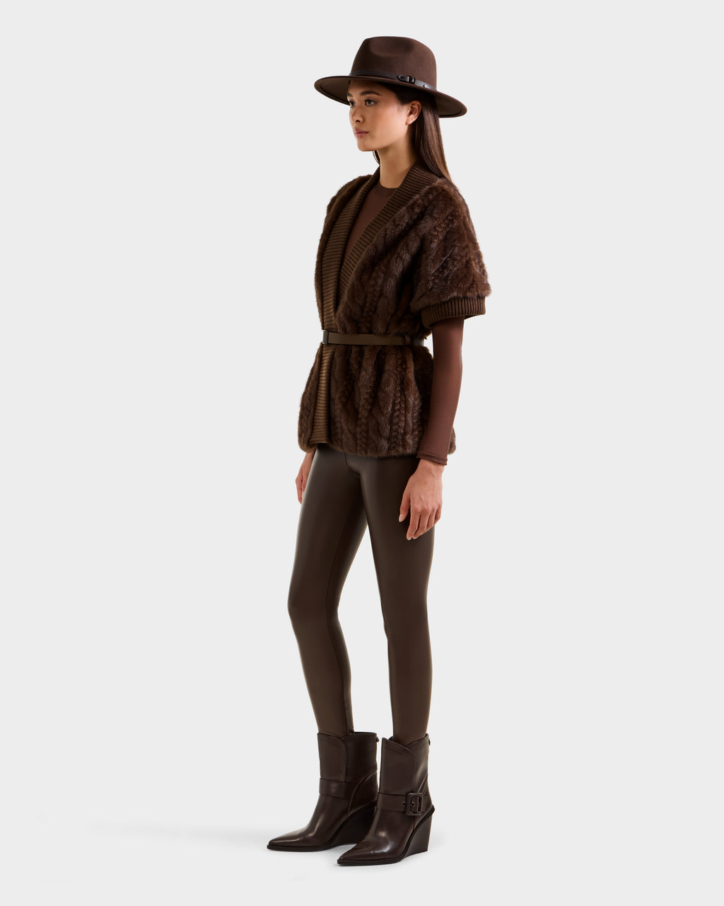 A woman in a stylish Rudsak MENDOTA F - 8525561 Mocha outfit, featuring a brown wide-brim hat, textured cardigan layered over a faux fur long-sleeve top, brown leggings, and heeled ankle boots stands against a plain light background.
