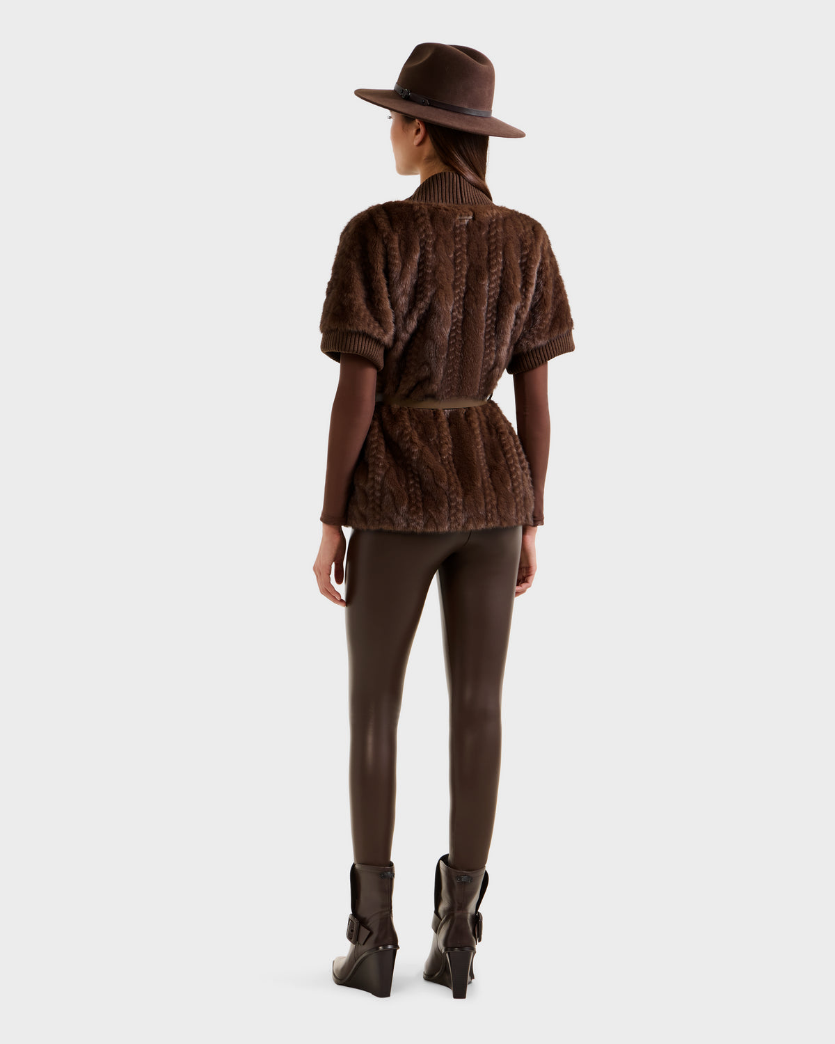 A woman faces away, wearing the Rudsak MENDOTA F - 8525561 Mocha faux fur top with short sleeves and a belt, paired with brown leggings and heeled ankle boots against a plain light background.
