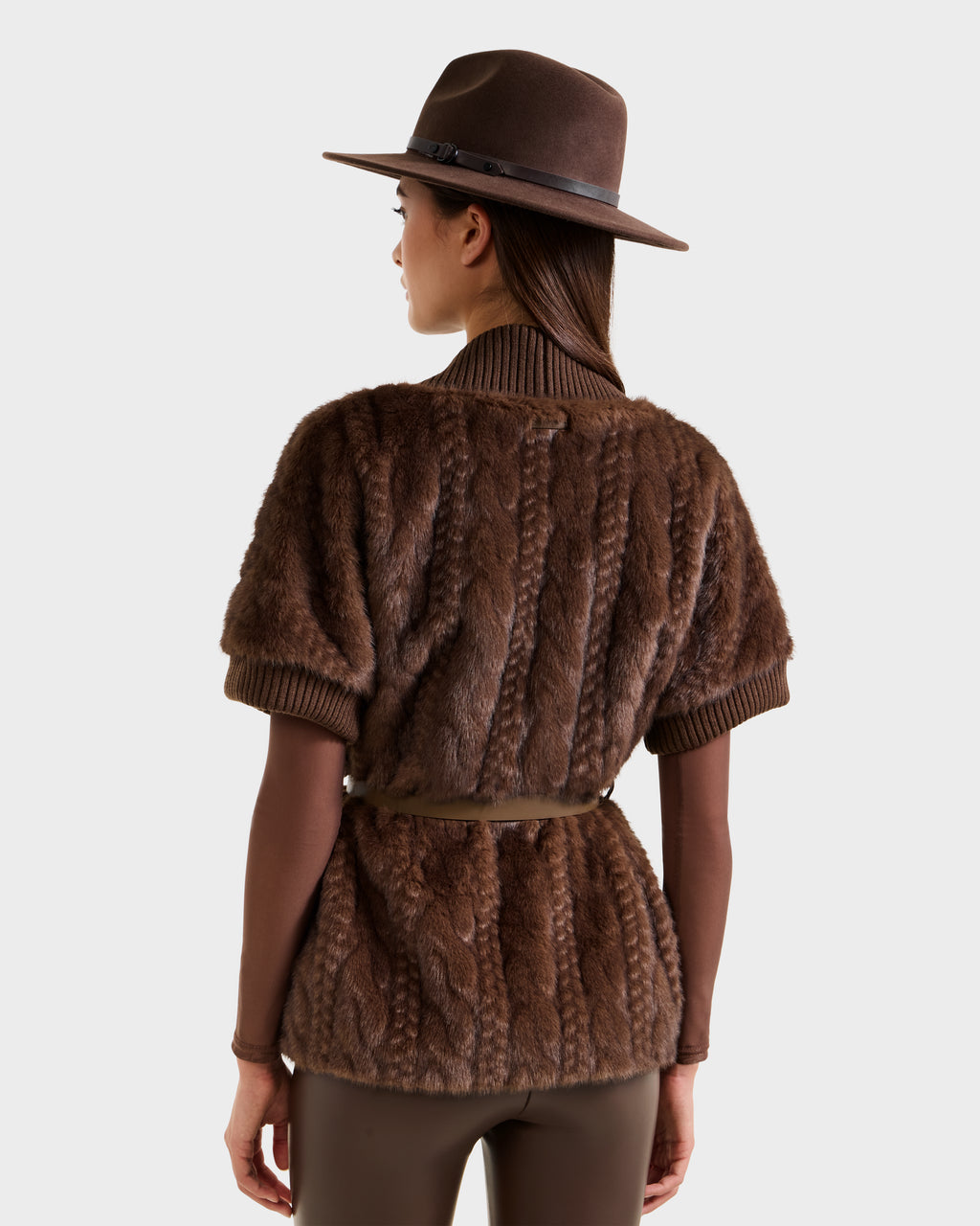 A woman is seen from behind against a plain background, wearing the Rudsak MENDOTA F - 8525561 Mocha short-sleeve belted faux fur jacket, a brown wide-brim hat, a brown long-sleeve shirt, and matching brown pants.
