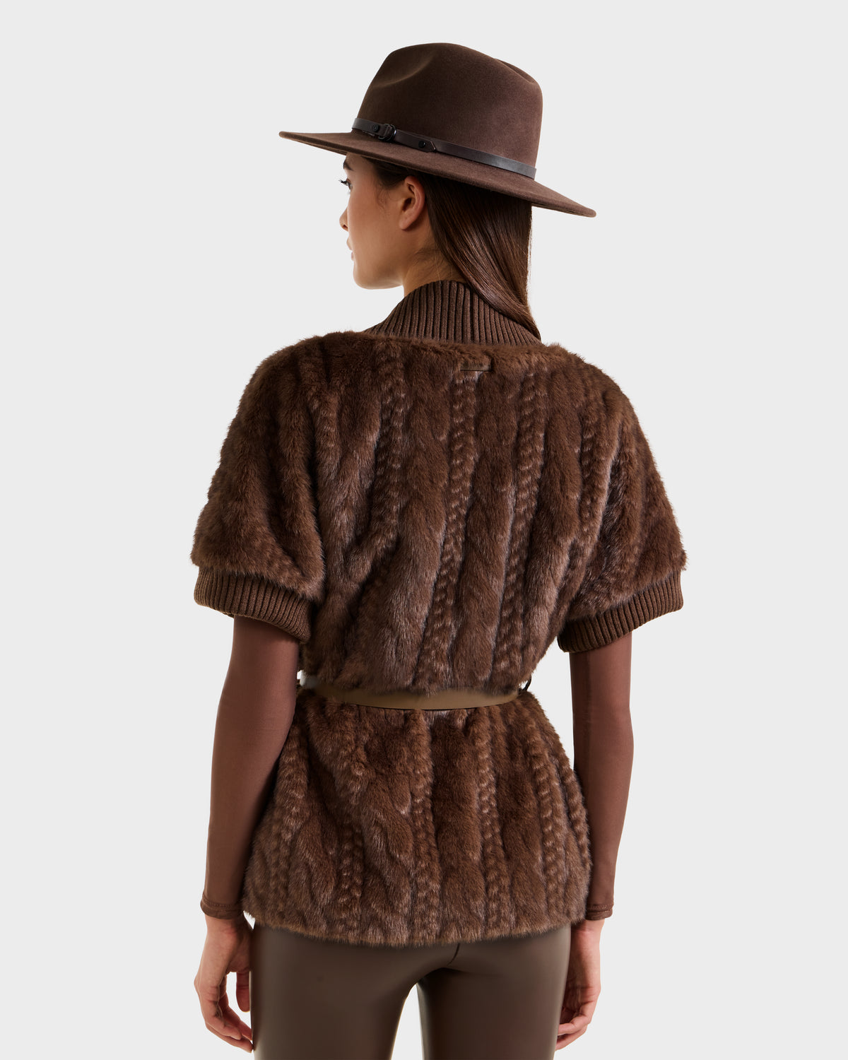 A woman is seen from behind against a plain background, wearing the Rudsak MENDOTA F - 8525561 Mocha short-sleeve belted faux fur jacket, a brown wide-brim hat, a brown long-sleeve shirt, and matching brown pants.