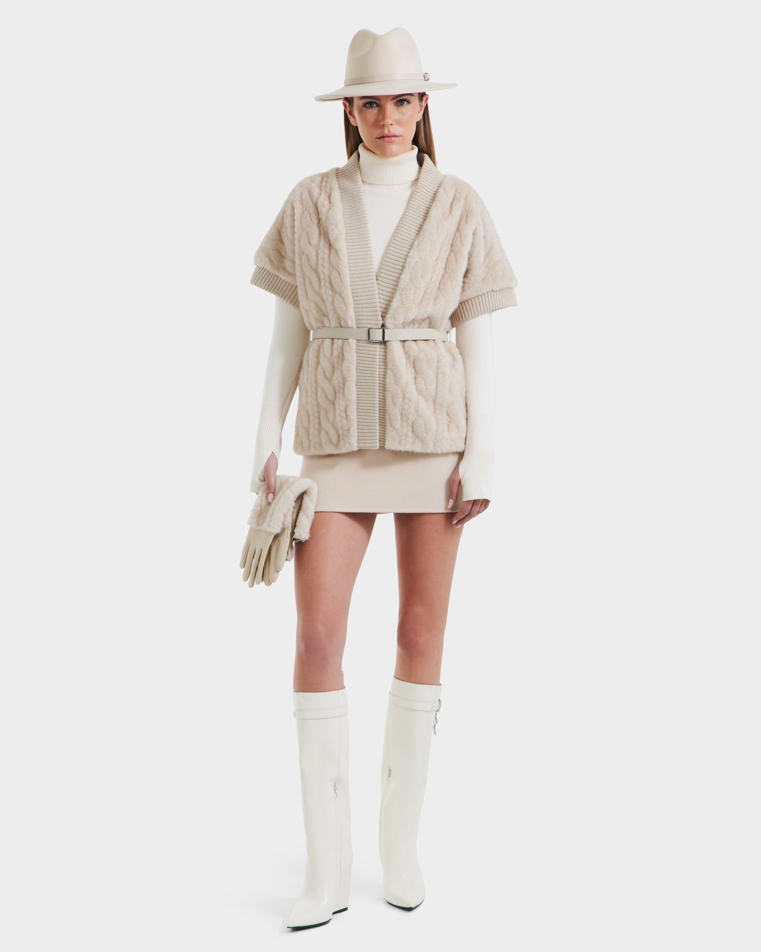 A woman models the Rudsak MENDOTA F - 8525561 Vanilla, pairing a cream hat, belted knit cardigan, wrap top, mini skirt, long gloves, knee-high white boots, and clutch in a chic beige and white palette inspired by Rudsak's signature style.