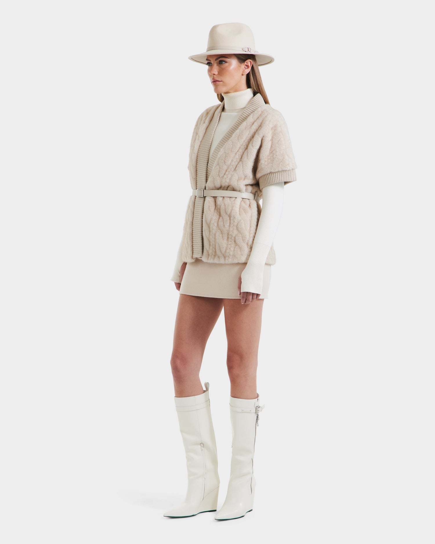 A woman models the Rudsak MENDOTA F - 8525561 Vanilla belted textured cardigan over a cream turtleneck, paired with a beige mini skirt and knee-high white boots, standing against a plain white background.
