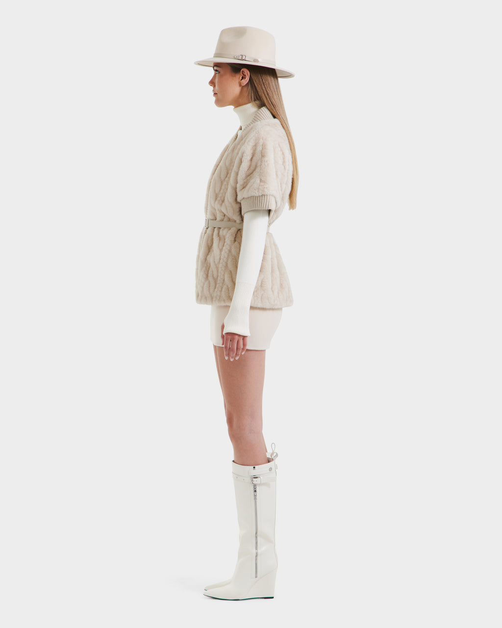 Woman in profile wears a beige hat, cream turtleneck, Rudsak MENDOTA F - 8525561 Vanilla textured short jacket with belt, cream shorts, and tall white wedge-heeled boots against a white background.
