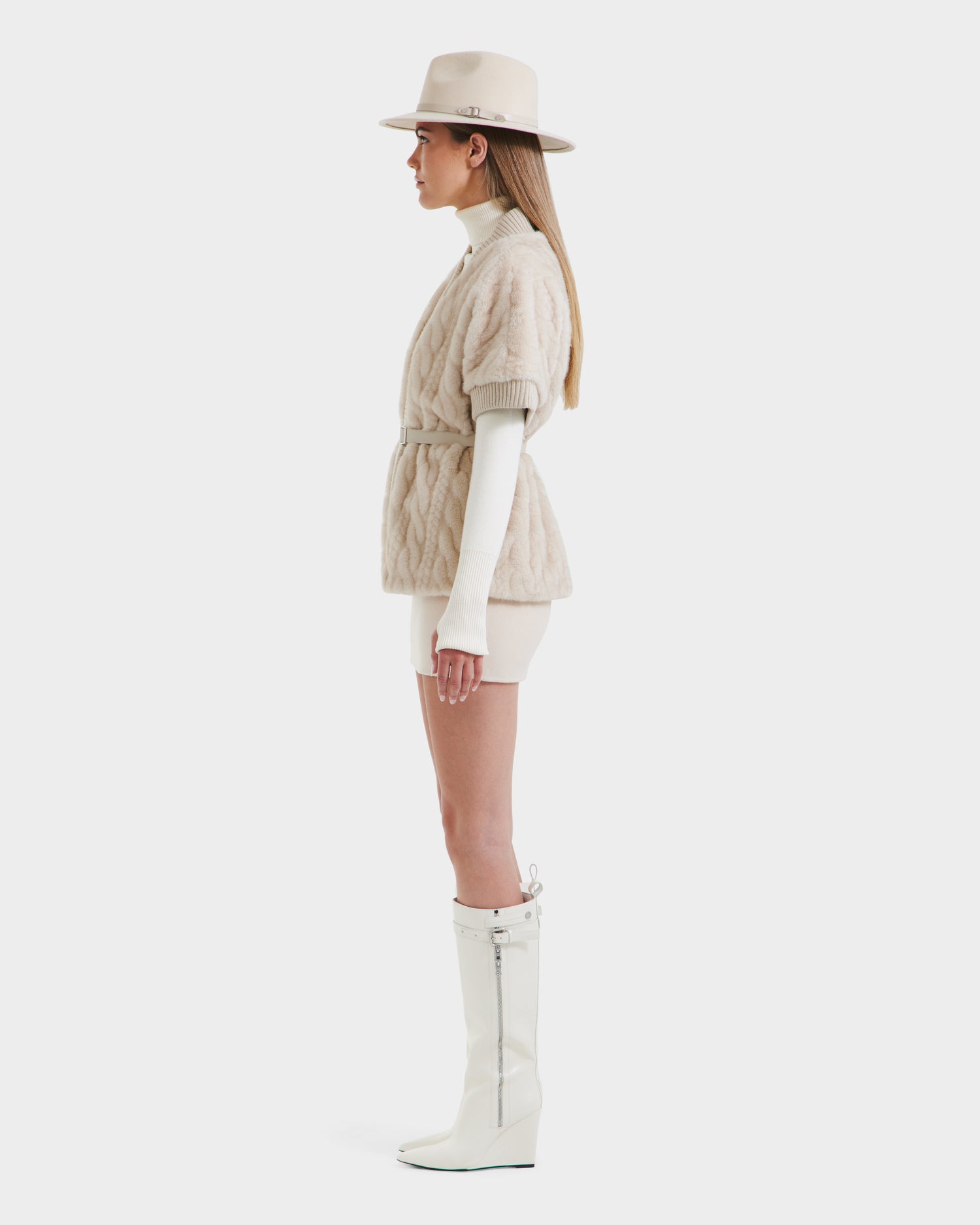 Woman in profile wears a beige hat, cream turtleneck, Rudsak MENDOTA F - 8525561 Vanilla textured short jacket with belt, cream shorts, and tall white wedge-heeled boots against a white background.