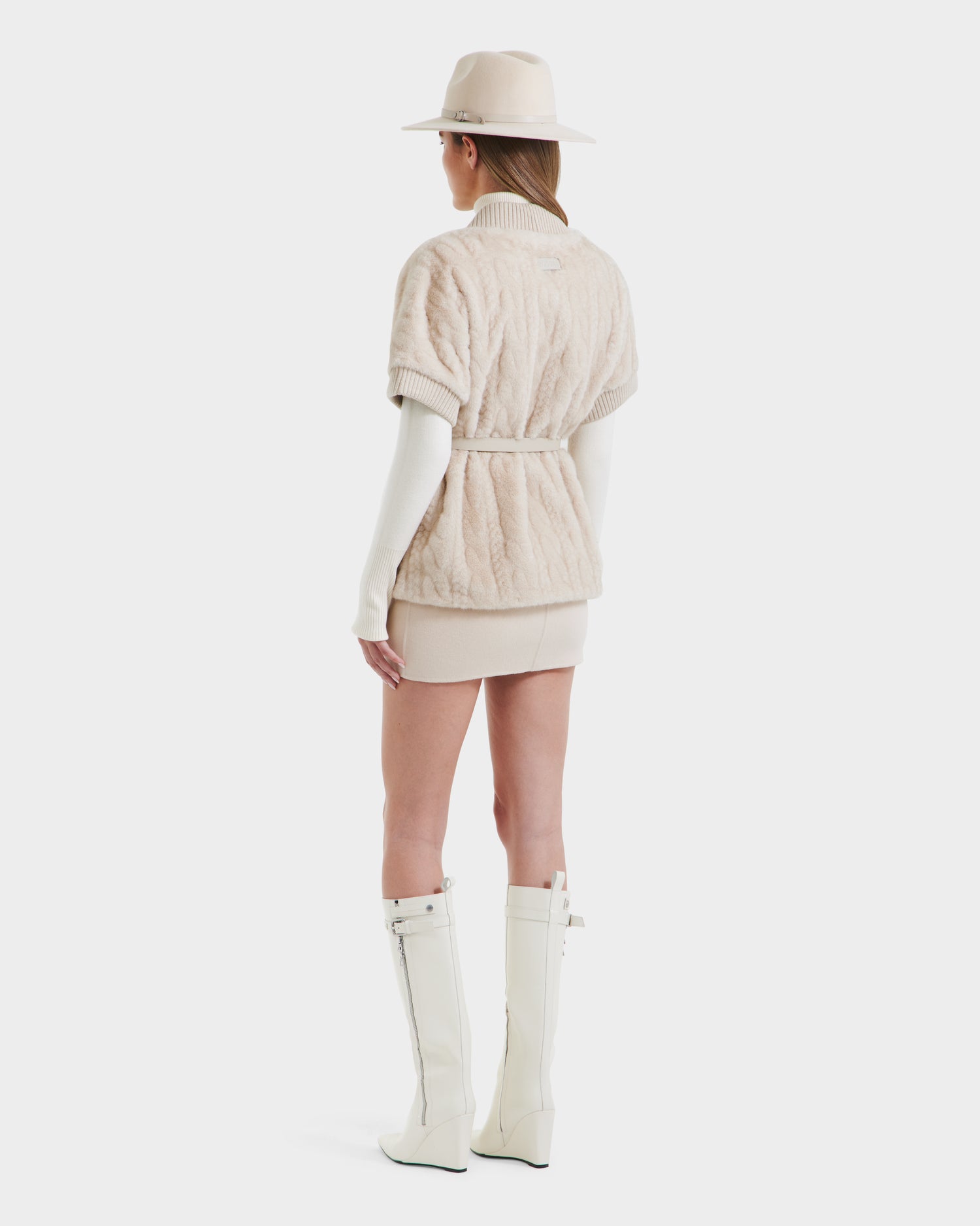 A woman faces away against a plain white background, wearing the Rudsak MENDOTA F - 8525561 Vanilla knee-high boots with zippers, styled with a light hat, belted textured jacket over a wrap top, and a cream mini skirt.