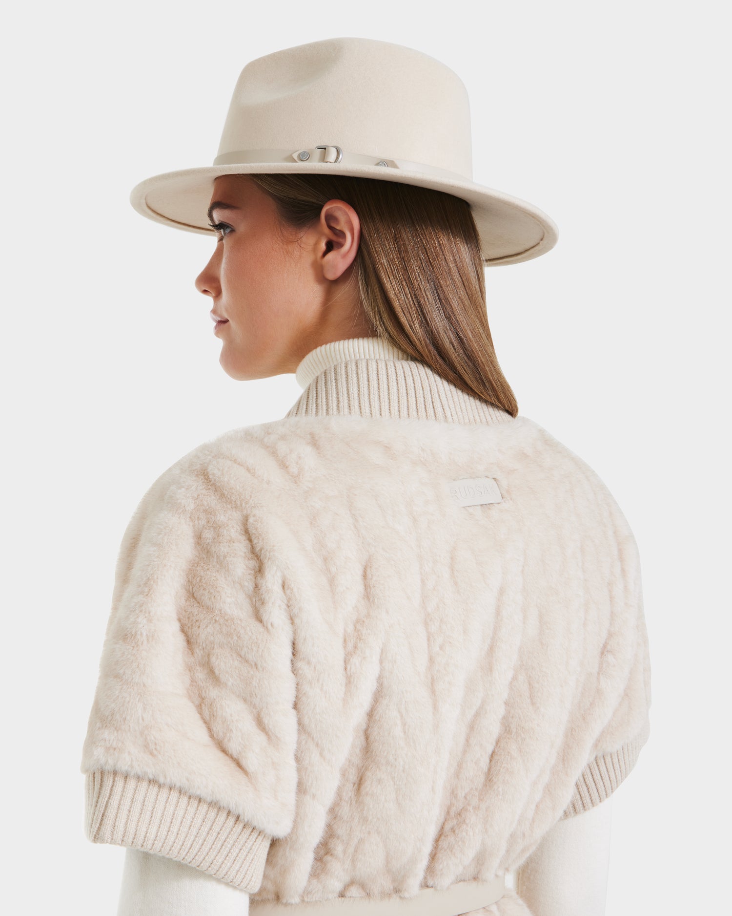 A woman with straight brown hair is shown from the side and back wearing a cream wide-brim hat and the Rudsak MENDOTA F - 8525561 Vanilla jacket over a ribbed turtleneck, set against a plain white background.