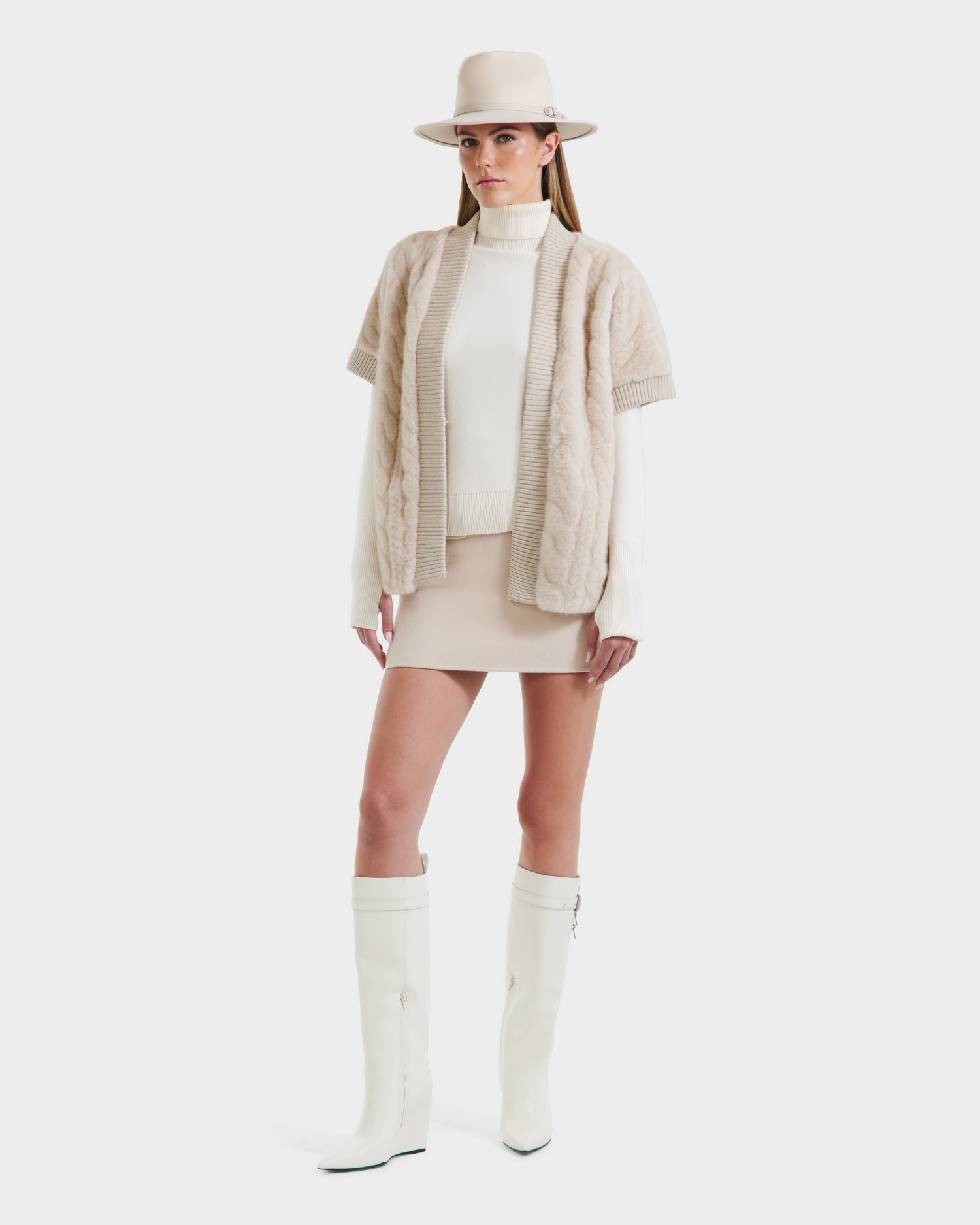 A woman wearing a beige hat, cream turtleneck, beige short-sleeve cardigan, Rudsak MENDOTA F - 8525561 Vanilla mini skirt, and knee-high white boots stands against a white background.