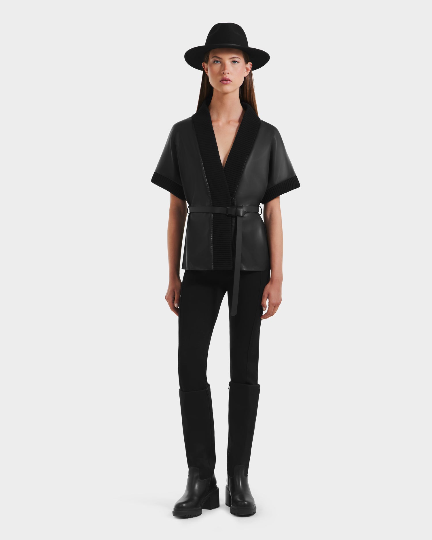 A woman faces forward in a black wide-brimmed hat, the Rudsak MENDOTA - 8525665 vegan leather wrap top with short sleeves and belt, black pants, and knee-high black boots against a plain white background.