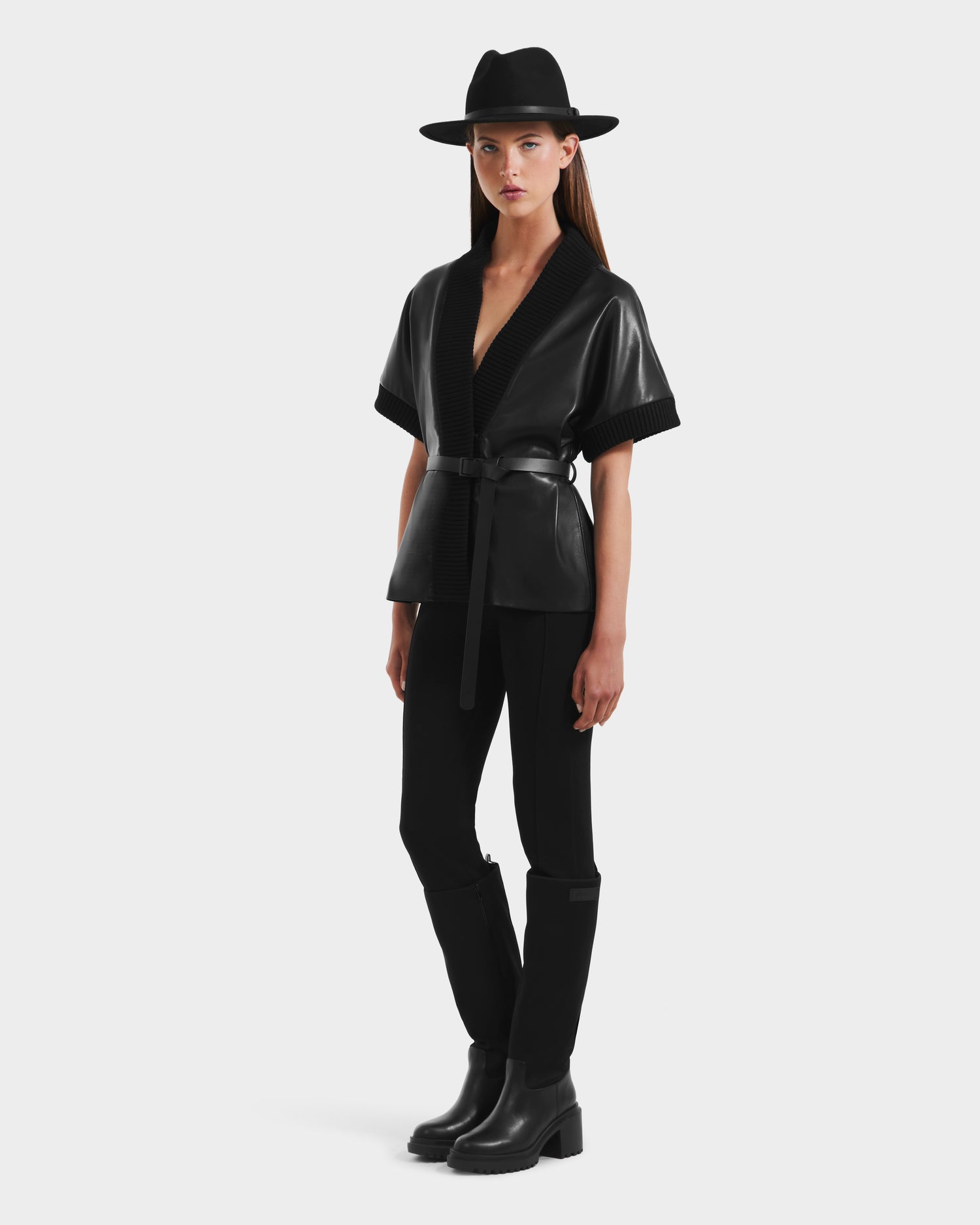 A woman stands against a plain white background wearing a wide-brimmed black hat, black pants, black boots, and the Rudsak MENDOTA - 8525665 Black vegan leather wrap top with short sleeves.