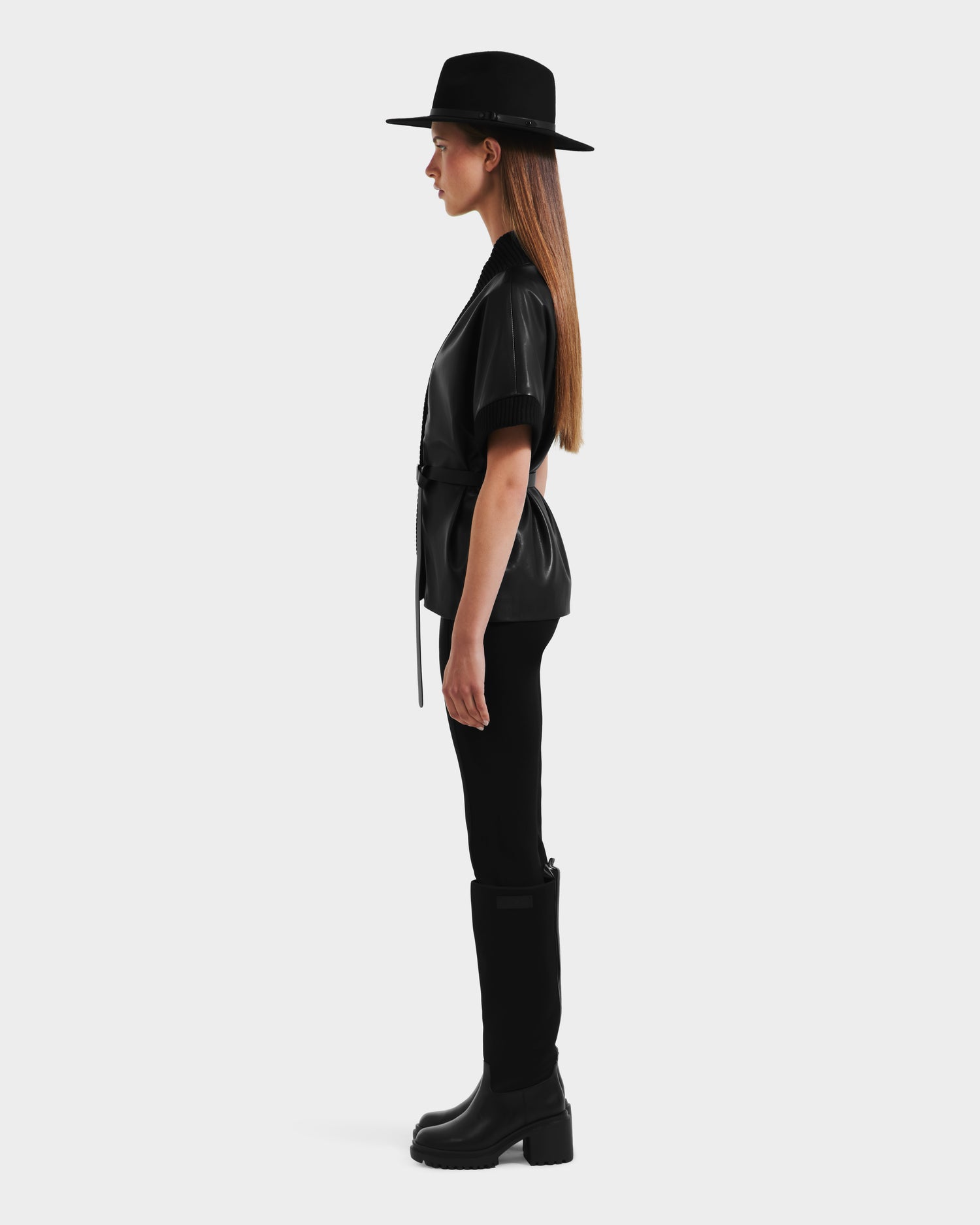A woman in profile wears a wide-brimmed black hat, the Rudsak MENDOTA - 8525665 Black vegan leather wrap top with short sleeves, black pants, and tall black boots against a plain white background.