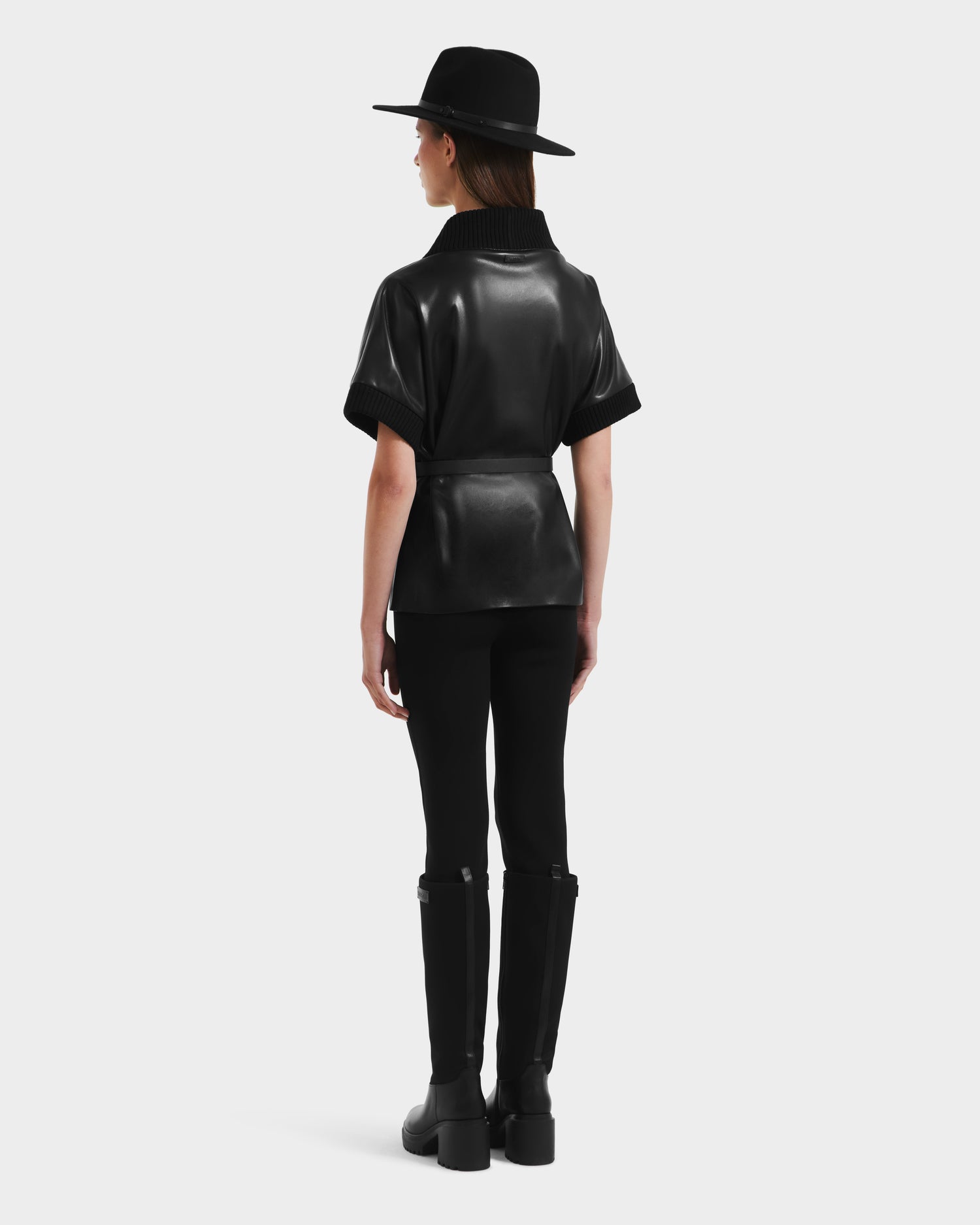 A person stands facing away, wearing a wide-brim black hat, black pants, knee-high black boots, and the MENDOTA - 8525665 Black vegan leather wrap top by Rudsak with short sleeves against a plain light background.
