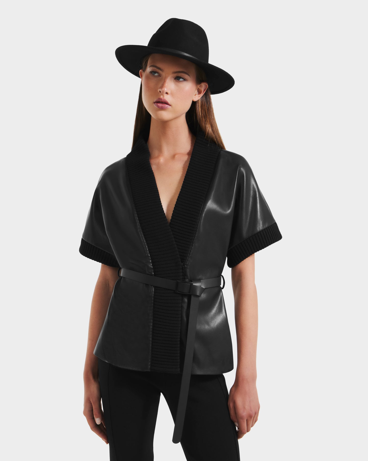 A woman in a black wide-brim hat, black pants, and the Rudsak MENDOTA - 8525665 Black vegan leather wrap top with textured collar and trim stands against a plain white background.