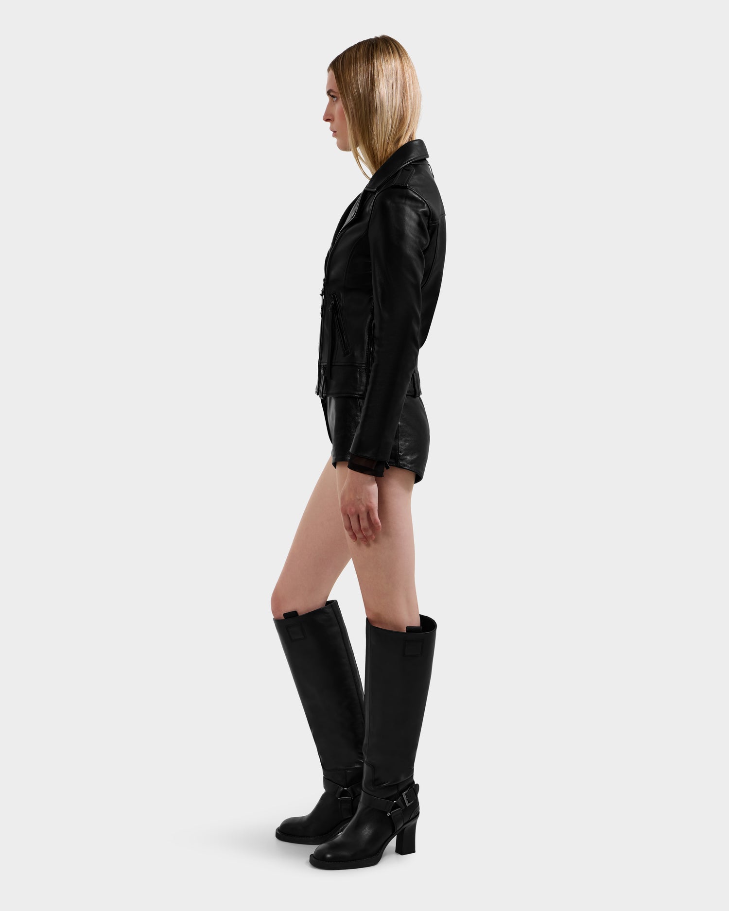 A woman with straight blonde hair stands in profile against a plain light gray background, wearing the MERGO - 8122009 BLACK biker jacket by Rudsak, paired with black shorts and knee-high black boots.