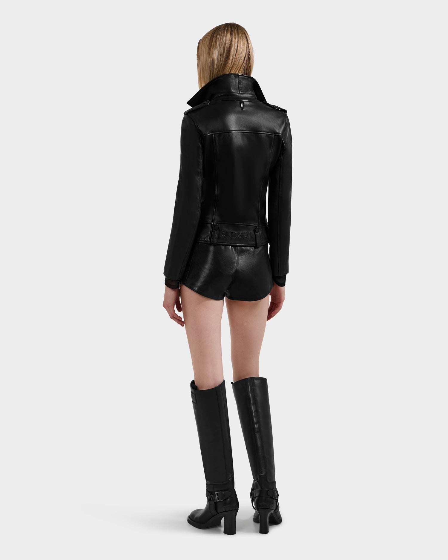 A woman with long, straight hair faces away, wearing the Rudsak MERGO - 8122009 BLACK lambskin leather biker jacket, black shorts, and tall heeled black boots against a plain light gray background.