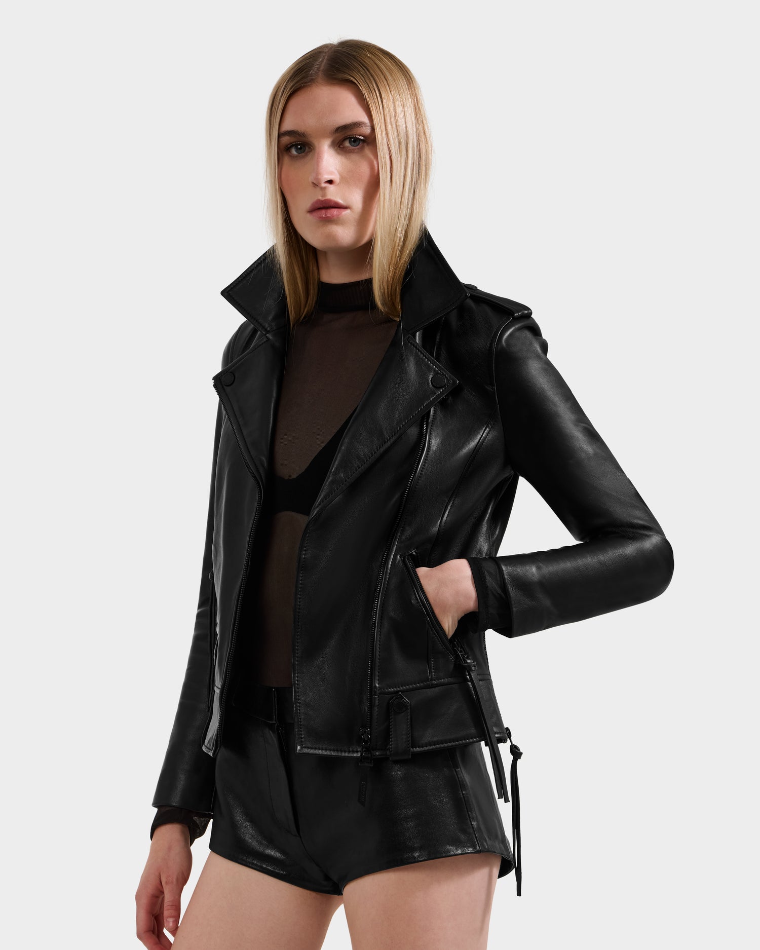 A person with long, straight blonde hair wears the Rudsak MERGO - 8122009 BLACK women's biker jacket and matching black leather shorts over a sheer dark top, standing against a plain light gray background.