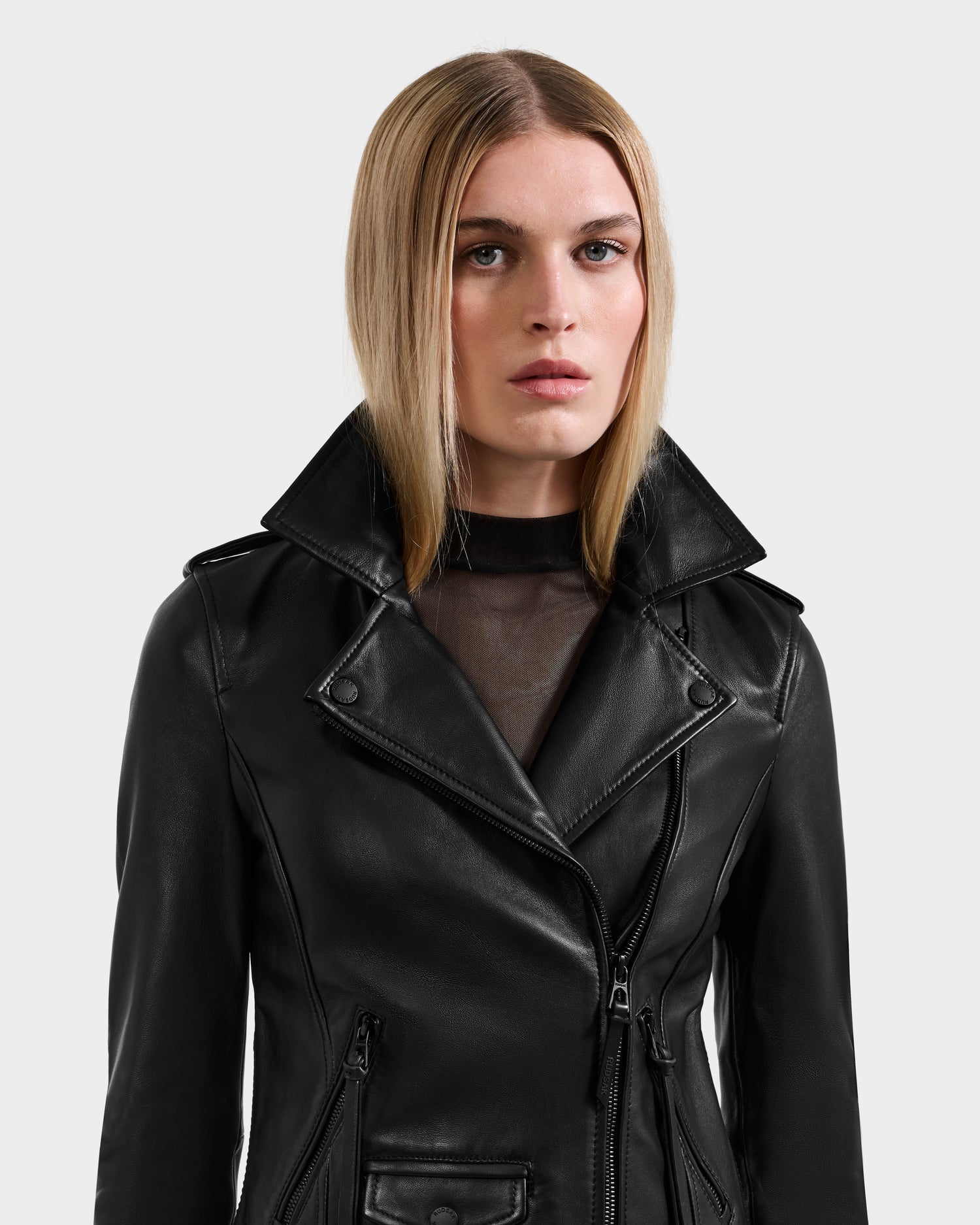 A woman with straight, blonde hair wears the Rudsak MERGO - 8122009 BLACK lambskin leather biker jacket with a high collar and zippers over a sheer black top, standing against a plain light background.