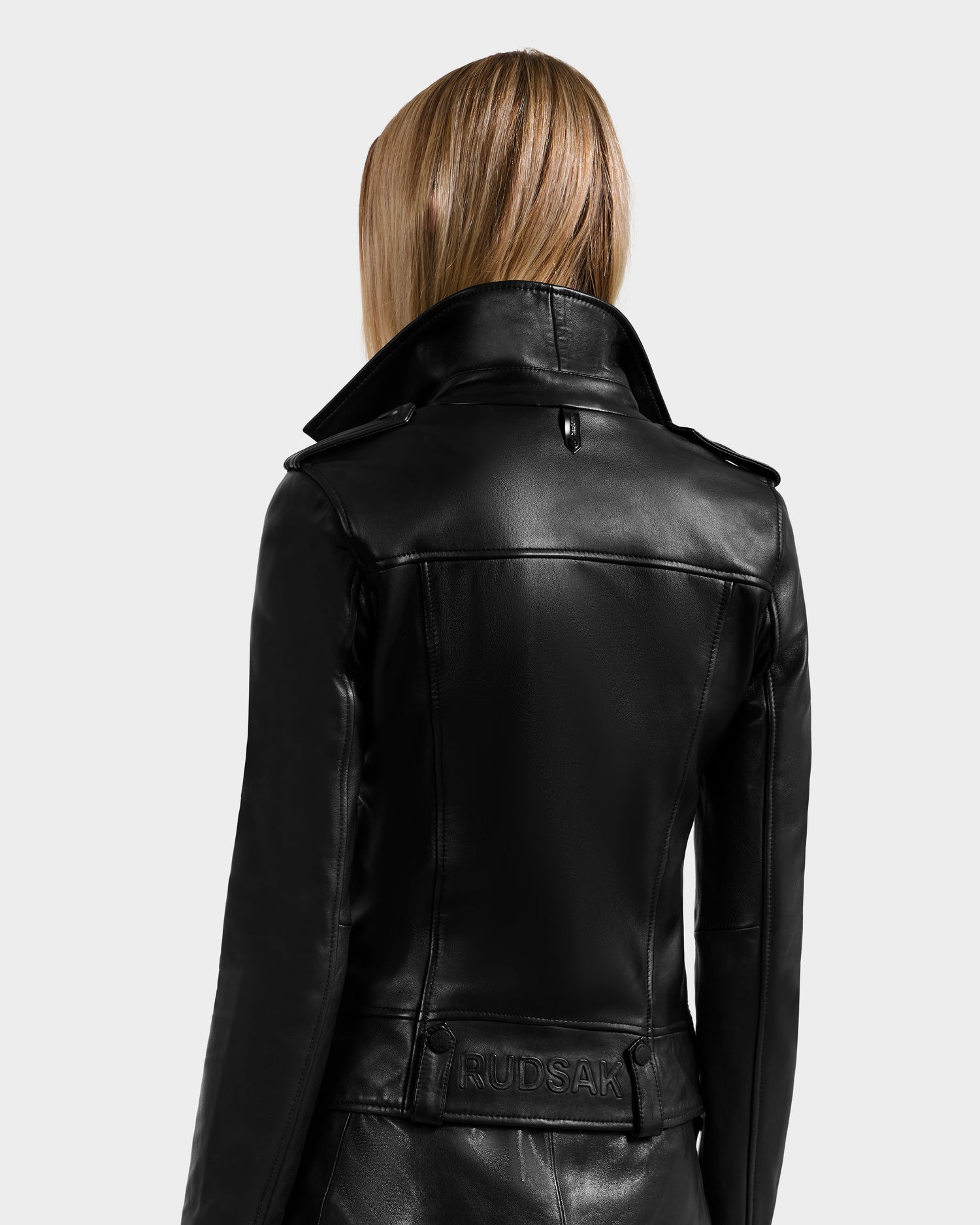 A woman with straight, light brown hair is shown from behind, wearing the fitted black Rudsak MERGO - 8122009 lambskin leather biker jacket featuring a turned-up collar and an embossed RUDSAK patch above the lower back.