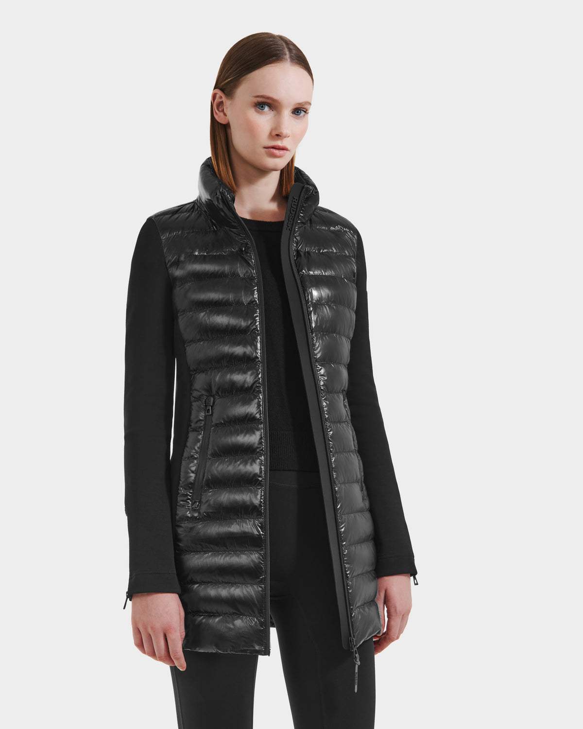 A woman with straight brown hair wears the Rudsak MERINA C - 8125648 Black long quilted down jacket with fabric sleeves and zipper details, paired with black pants, against a plain light background.