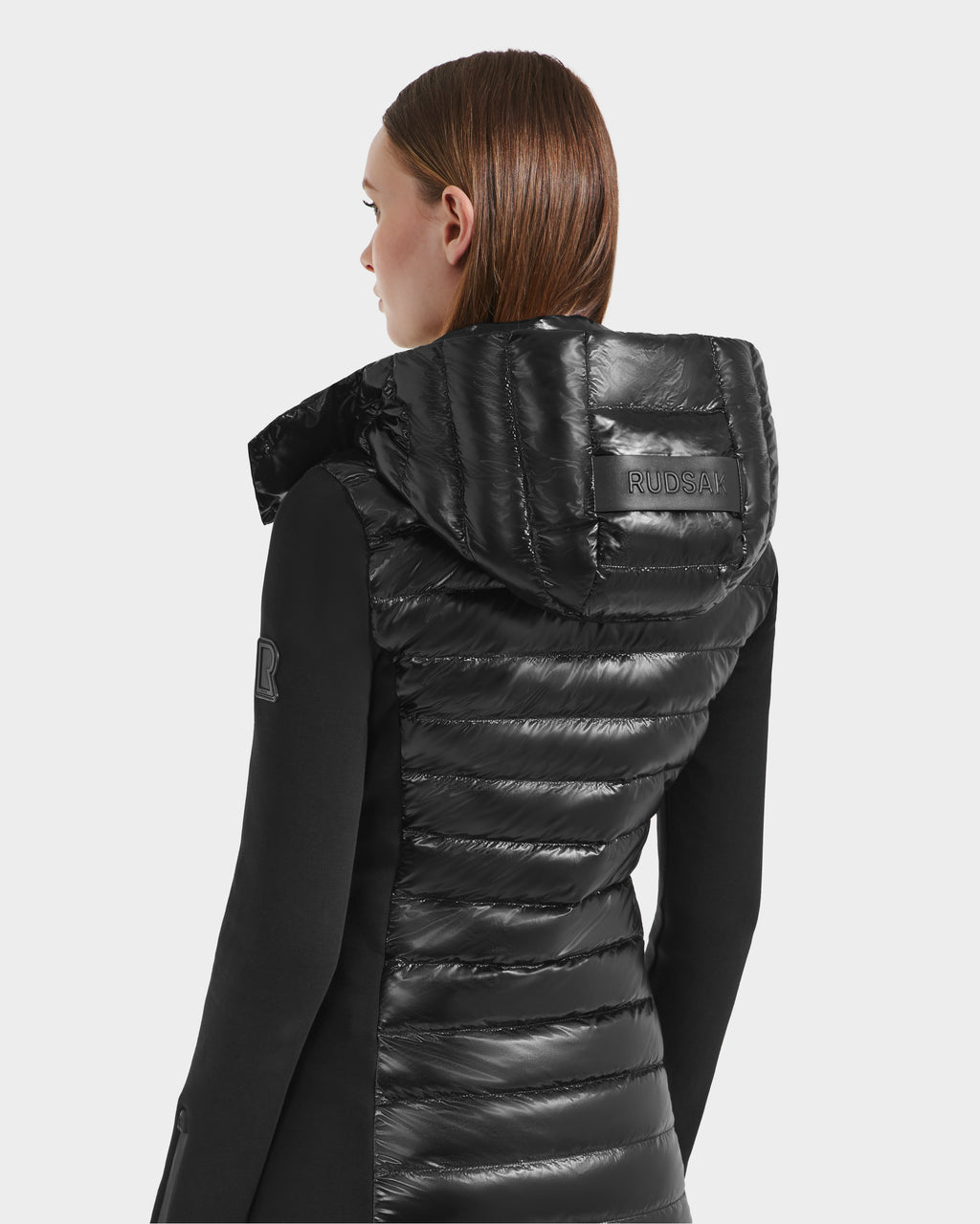 A woman with straight brown hair faces away, wearing the Rudsak MERINA C - 8125648 Black women’s light jacket with a hood featuring RUDSAK embossed on it. She stands against a plain white background.
