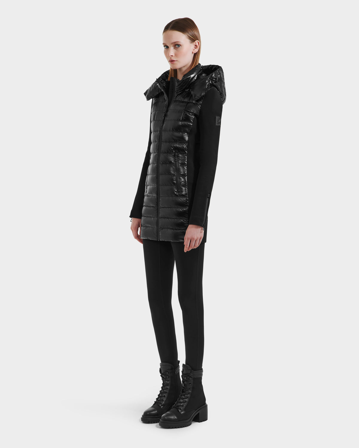 A woman stands facing forward in the Rudsak MERINA C - 8125648 Black quilted down jacket over a black long-sleeve top, black leggings, and black lace-up boots against a plain white background.