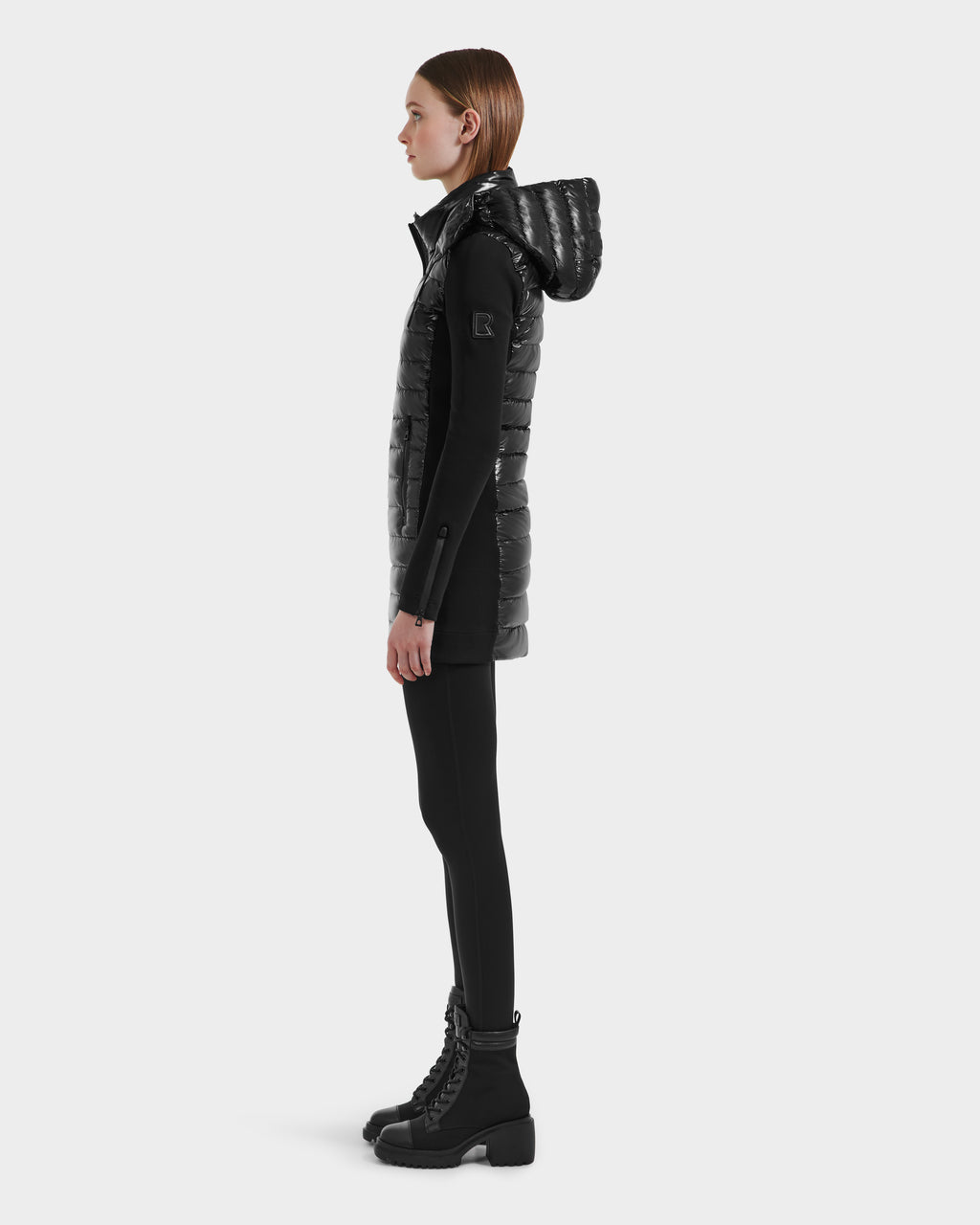 A woman stands in profile against a plain light background, wearing the Rudsak MERINA C - 8125648 Black hooded, quilted down long vest over black leggings and chunky black lace-up boots.