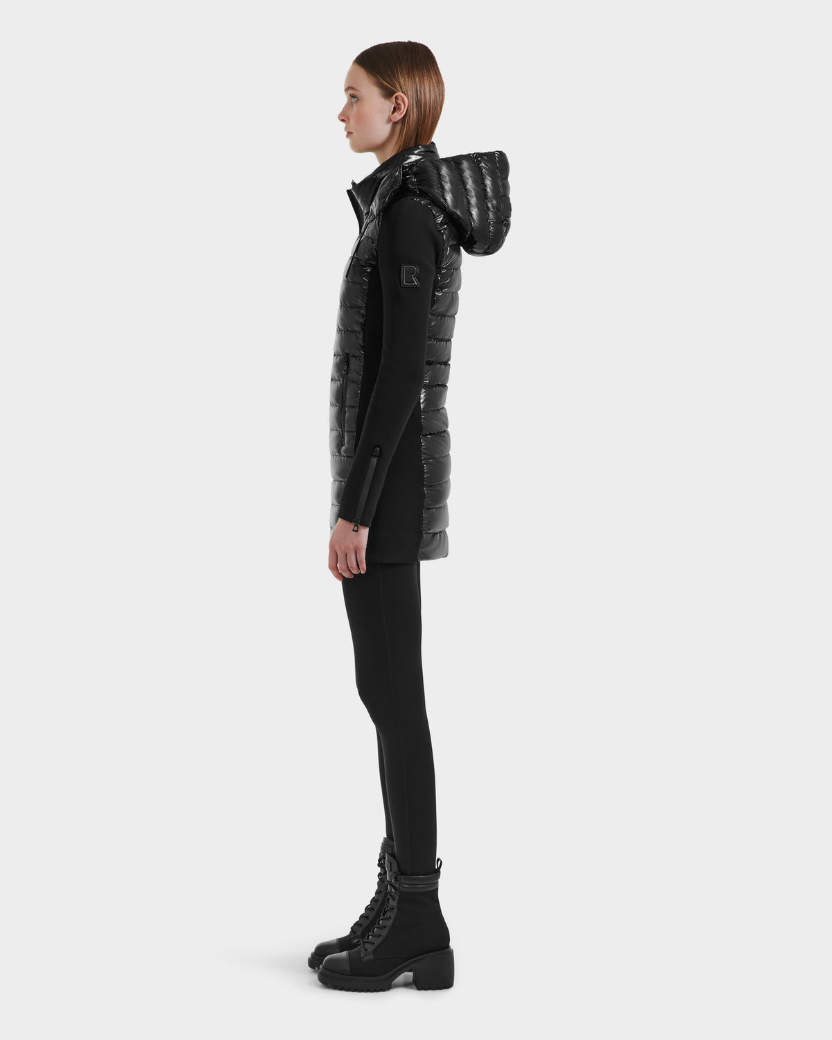 A woman stands in profile against a plain light background, wearing the Rudsak MERINA C - 8125648 Black hooded, quilted down long vest over black leggings and chunky black lace-up boots.