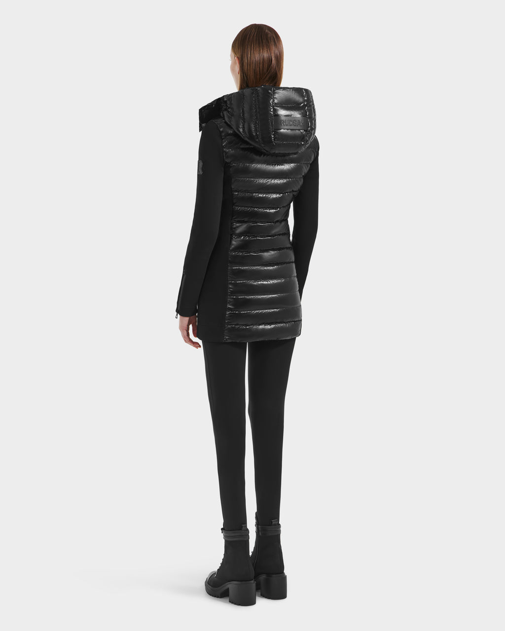 A person with straight brown hair faces away, wearing the Rudsak MERINA C - 8125648 Black long quilted down jacket with a glossy hood, black leggings, and black boots against a plain white background.