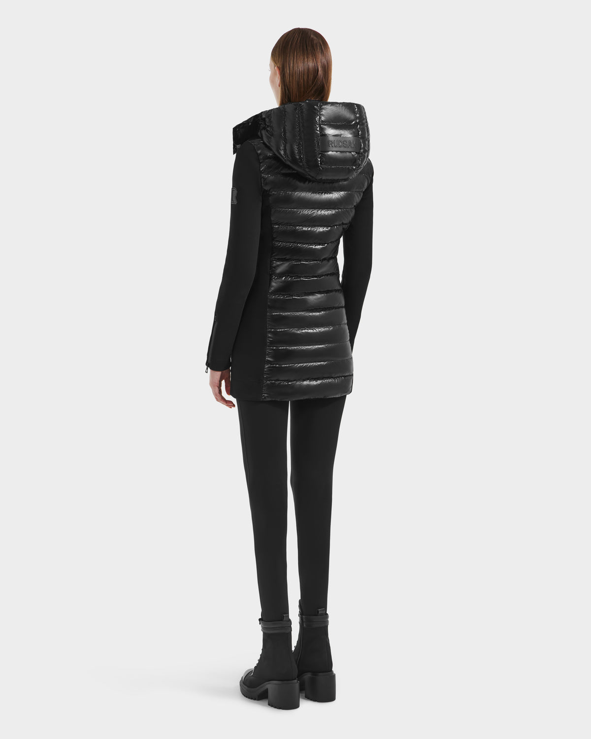A person with straight brown hair faces away, wearing the Rudsak MERINA C - 8125648 Black long quilted down jacket with a glossy hood, black leggings, and black boots against a plain white background.