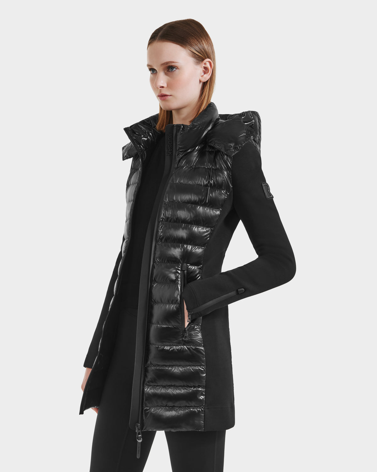 A woman with straight, light brown hair wears the Rudsak MERINA C - 8125648 Black long women’s jacket, featuring quilted, shiny sections on the front and sides. She stands facing left against a plain light background.