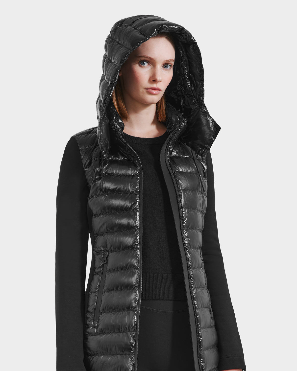 A woman with straight red hair wears the Rudsak MERINA C - 8125648 Black shiny lightweight hooded jacket over a long-sleeve black top, standing against a plain white background.
