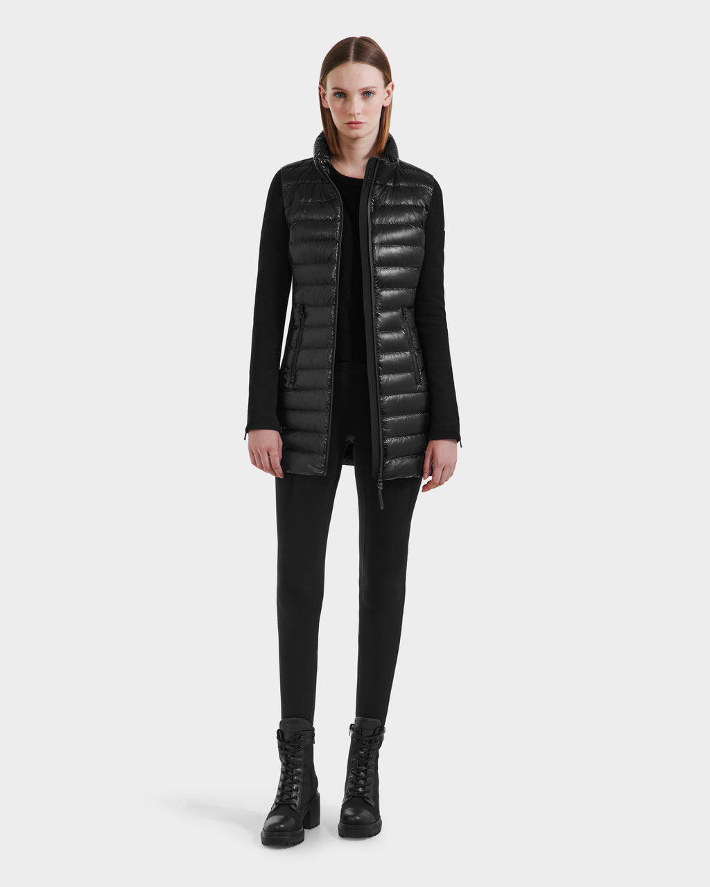 A person stands facing forward, wearing the MERINA C - 8125648 Black quilted down jacket vest by Rudsak over a black top, black pants, and black lace-up boots against a plain light background.