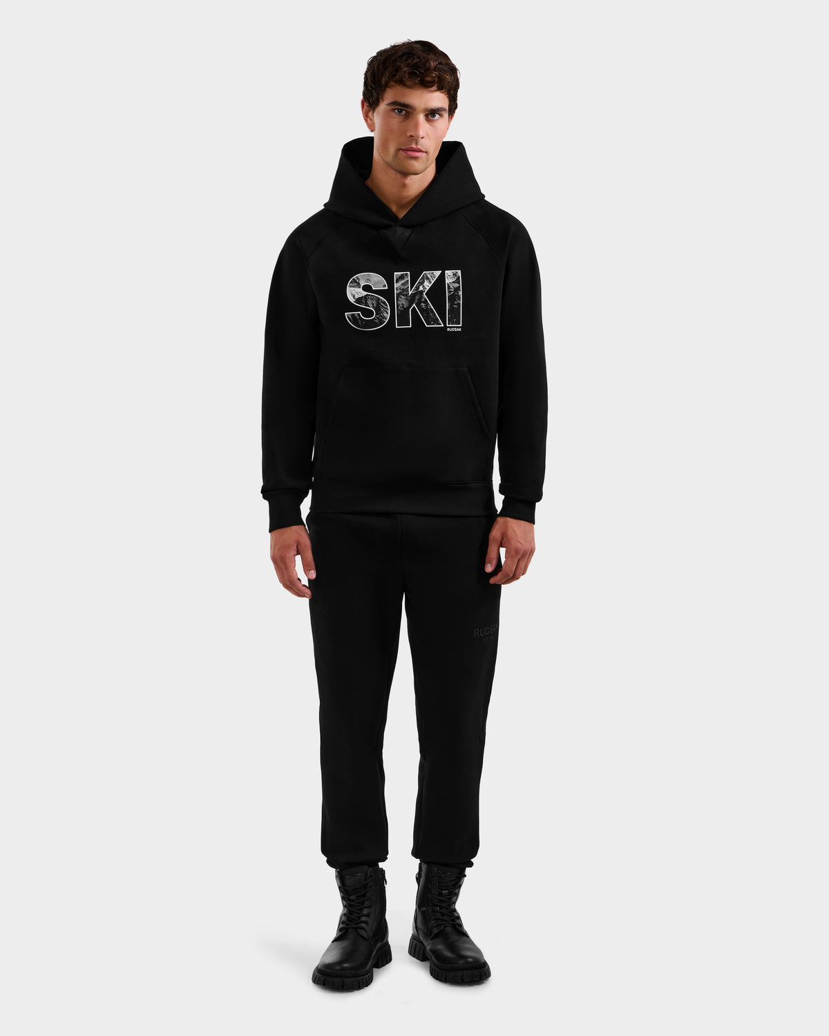 A man faces forward wearing the Rudsak MIKAEL - 6525647 Black premium cotton men’s ski hoodie with “SKI” on the chest, paired with black pants and boots, set against a plain white background.