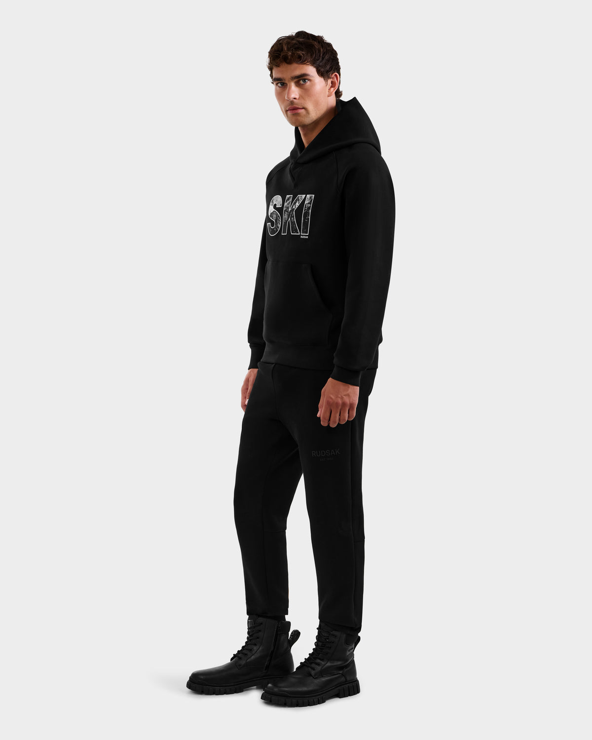 A man wearing the Rudsak MIKAEL - 6525647 Black premium cotton men’s ski hoodie with SKI lettering, black jogger pants, and lace-up boots stands against a plain background, facing slightly left with a neutral expression.