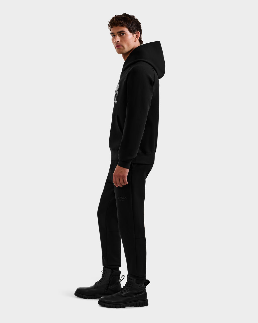 A man stands in profile against a plain white background, wearing the Rudsak MIKAEL - 6525647 Black premium cotton hoodie, matching black jogger pants, and black boots.