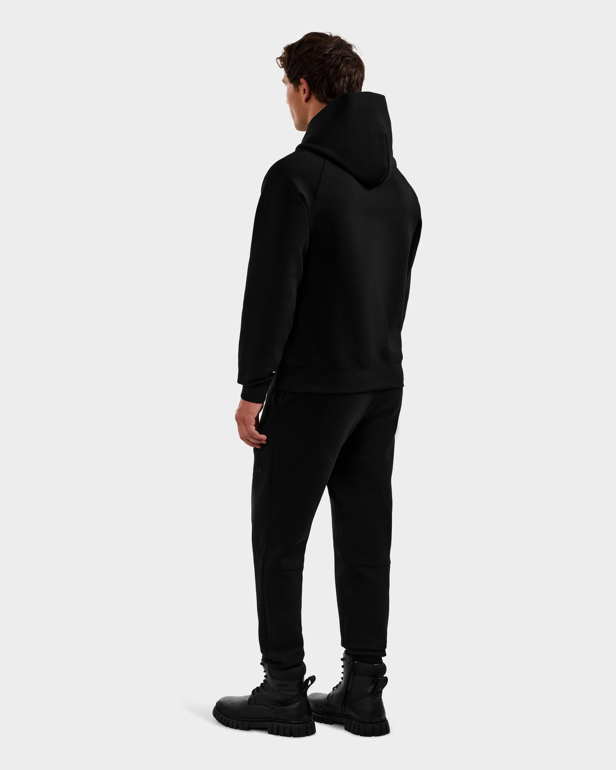 A person stands facing away, wearing the Rudsak MIKAEL - 6525647 Black men’s premium cotton ski hoodie, black jogger pants, and chunky boots against a plain light gray background.